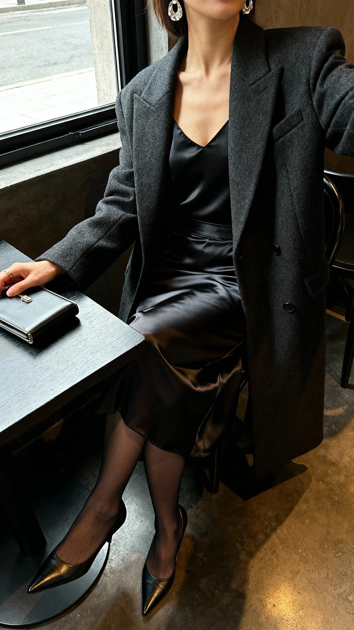 iPhone seated table selfie cropped at shoulders showing a woman in an evening look: black satin camisole under a charcoal wool tailored coat, high-waist satin midi skirt, sheer tights, pointed leather heels, and a slim clutch with statement earrings, face not visible, dim cafe lighting by window, natural iPhone photo quality.
