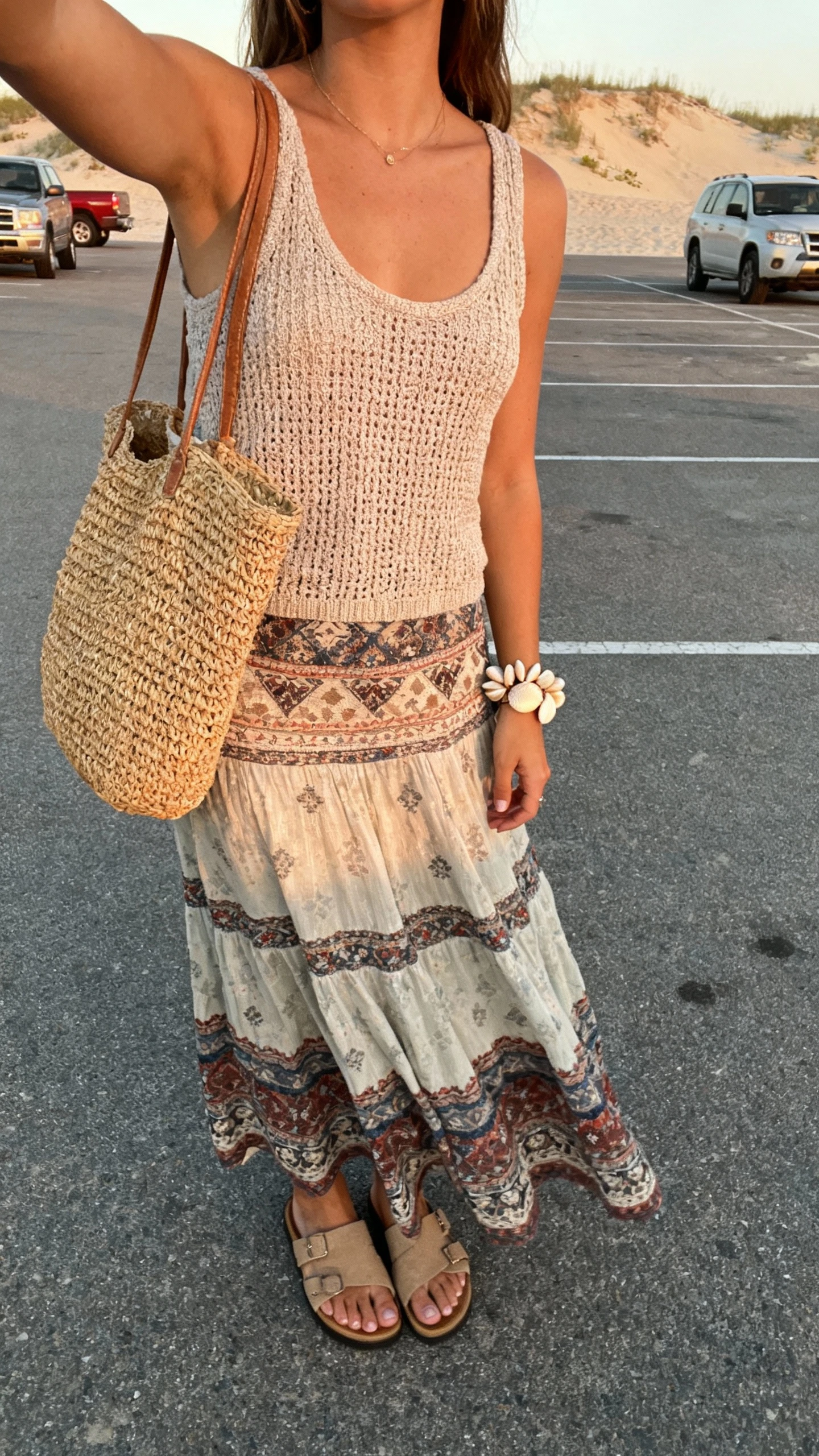 iPhone selfie cropped at neck showing a woman wearing a long boho skirt with a knit tank, slide sandals, straw tote, shell bracelet, face not visible, beach parking lot with dunes in background, soft afternoon light, natural iPhone photo quality