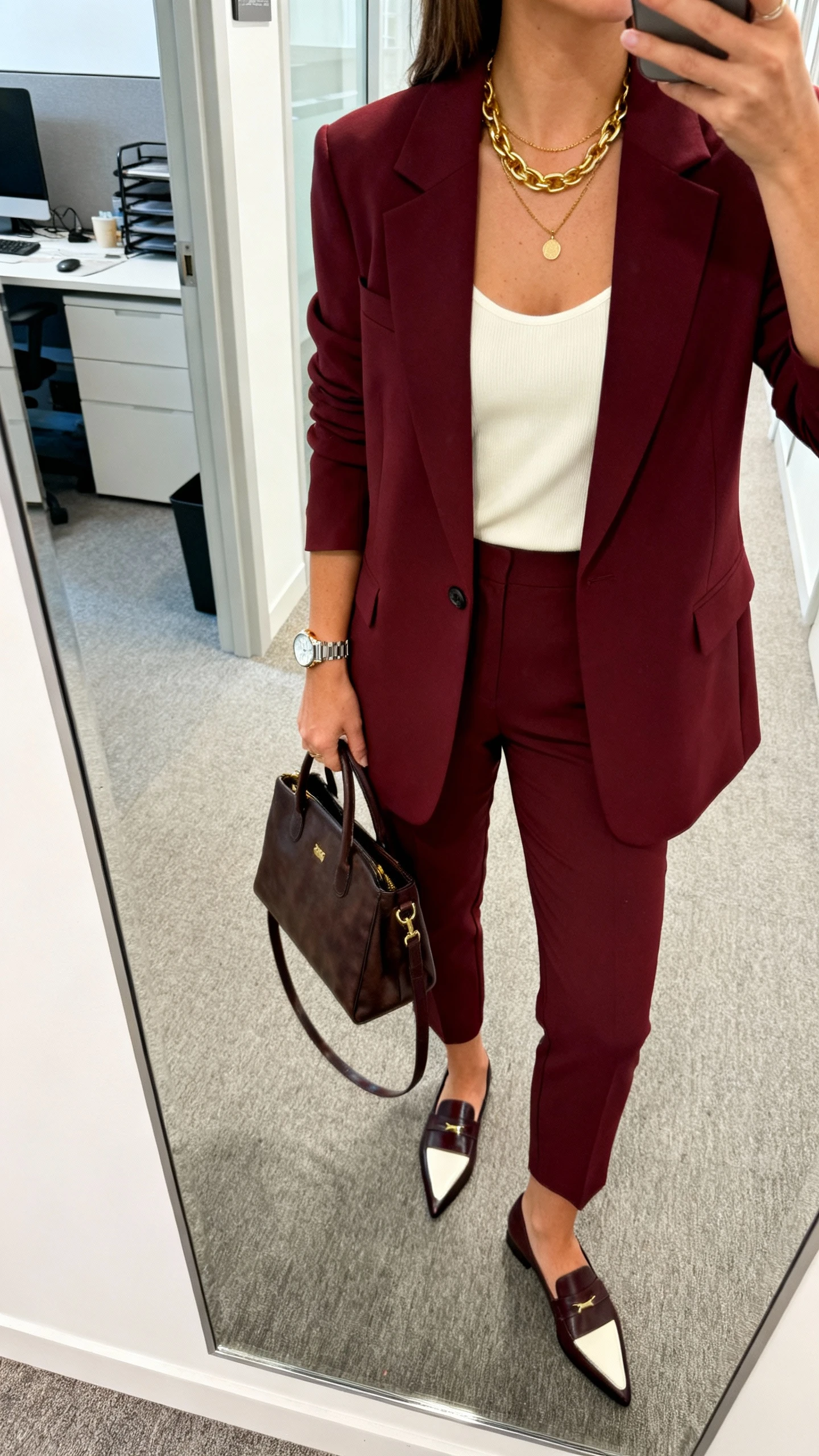iPhone selfie cropped at shoulders of a woman in a burgundy tailored blazer with matching trousers, cream camisole, pointed-toe loafers, layered gold necklace, structured tote and watch, face not visible, office hallway mirror, bright indoor lighting, natural iPhone aesthetic.