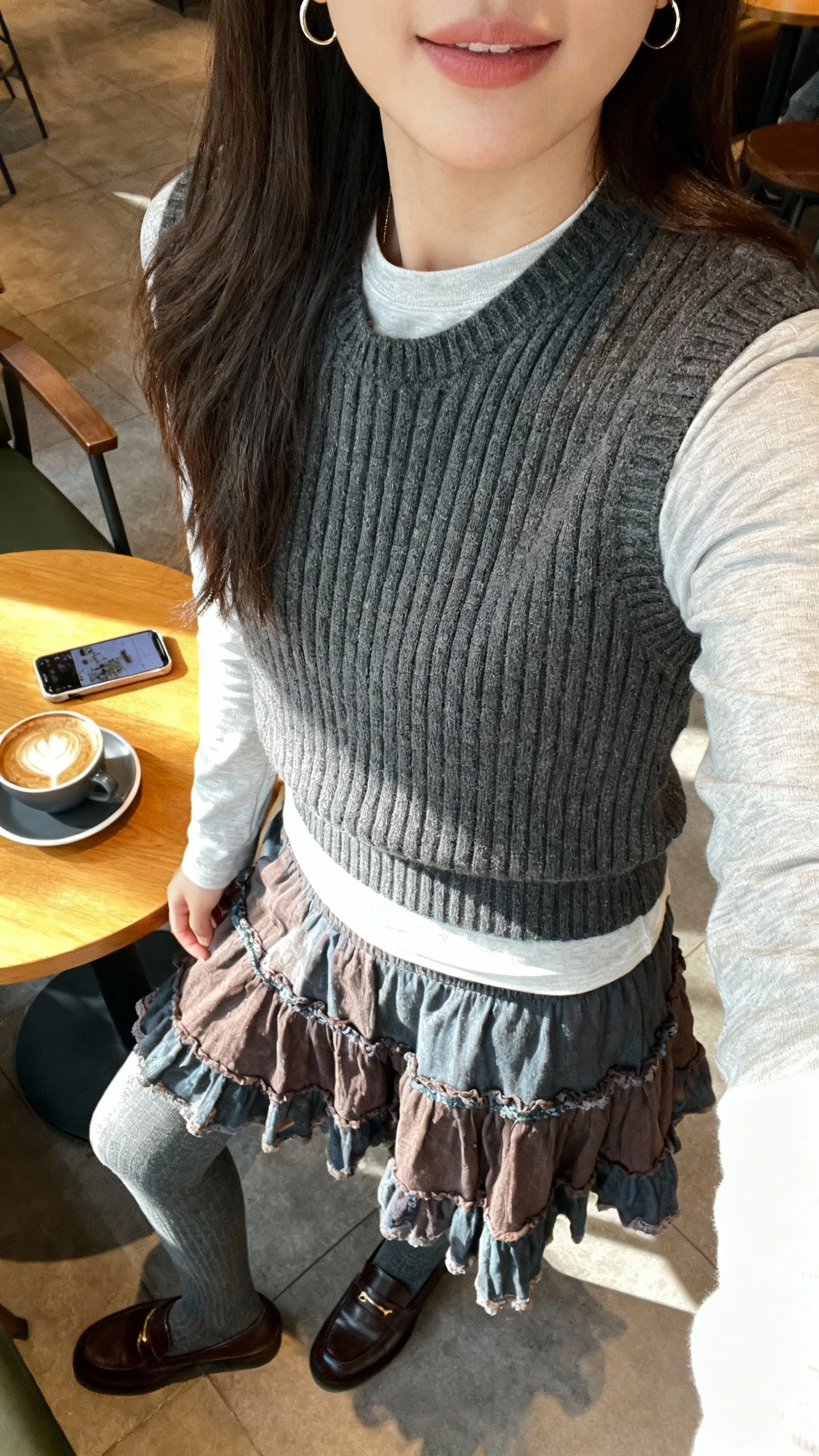 iPhone selfie cropped at shoulders showing a woman wearing a ribbed sweater vest over a long-sleeve tee paired with a tiered boho skirt, tights, loafers, and delicate hoop earrings, face not visible, cozy coffee shop table with morning light, natural iPhone photo quality.