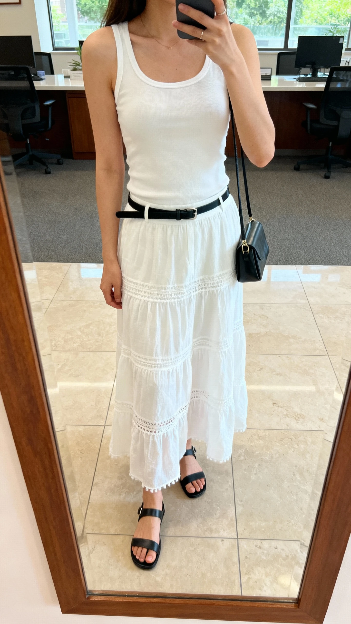iPhone selfie cropped at shoulders showing a woman wearing a white boho skirt, fitted white tank, sleek minimalist black sandals, thin black belt, small structured bag, face not visible, office lobby mirror, soft morning light, natural iPhone quality.