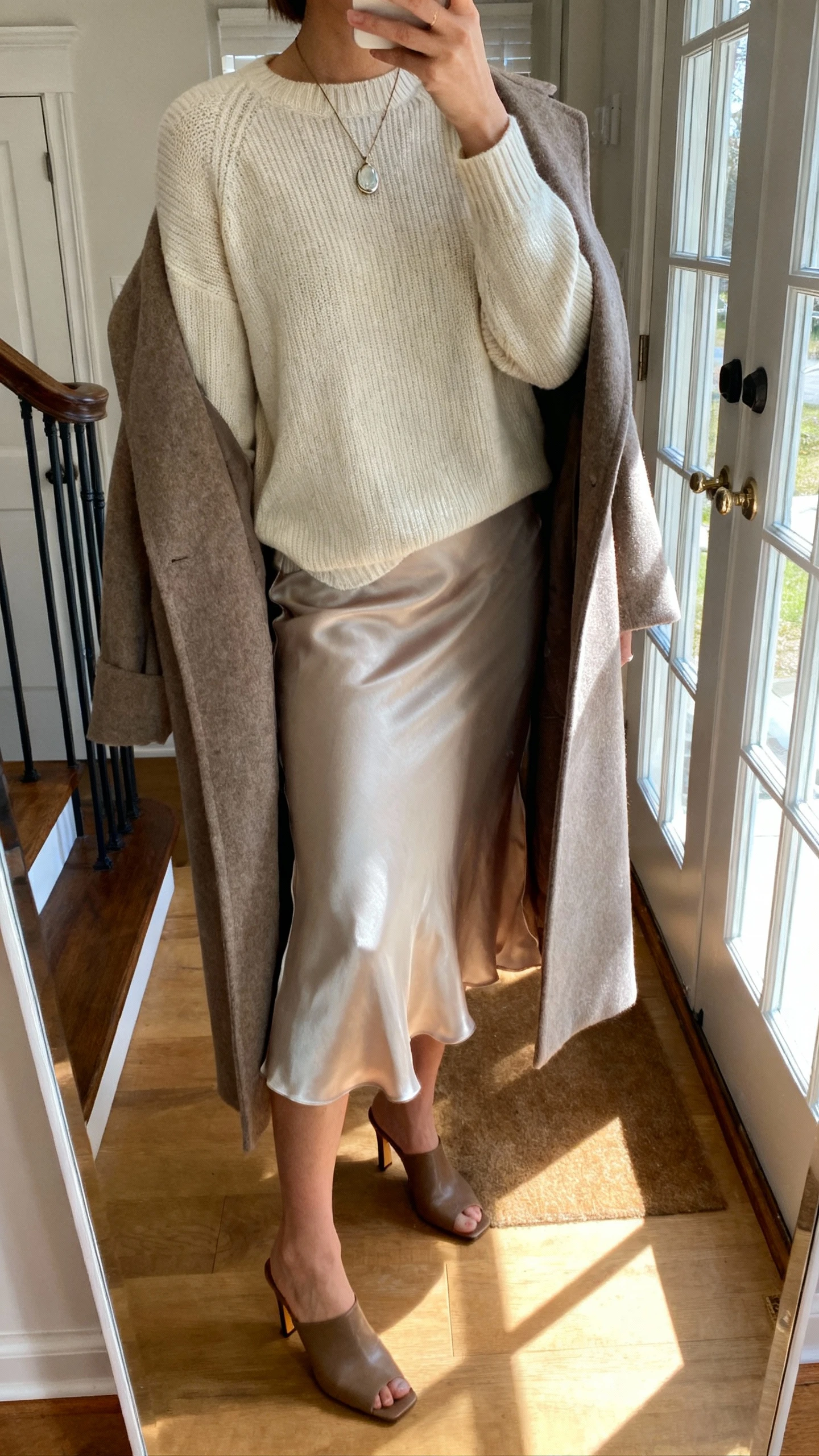iPhone selfie cropped at shoulders showing a woman layering a fine-gauge crewneck knit over a silky slip skirt, longline wool coat draped over shoulders, heeled mules, pendant necklace, face not visible, apartment entry mirror, afternoon window light, natural iPhone photo.