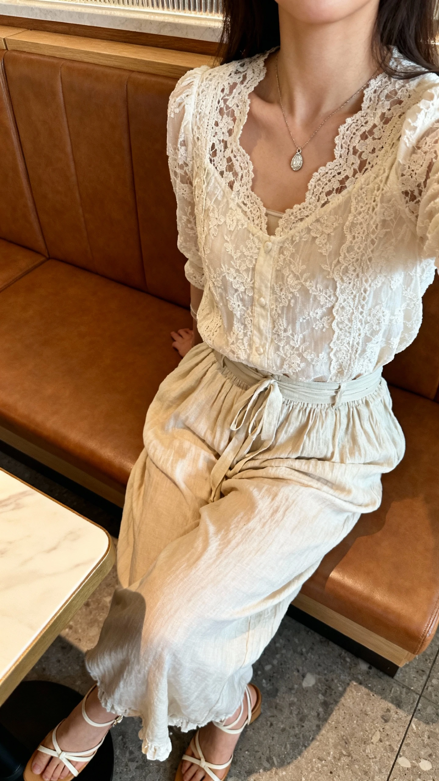 iPhone selfie cropped at shoulders showing a woman wearing a romantic lace top tucked into soft boho pants, dainty pendant necklace, strappy heels, face not visible, cozy restaurant banquette, warm ambient light, natural iPhone photo quality.