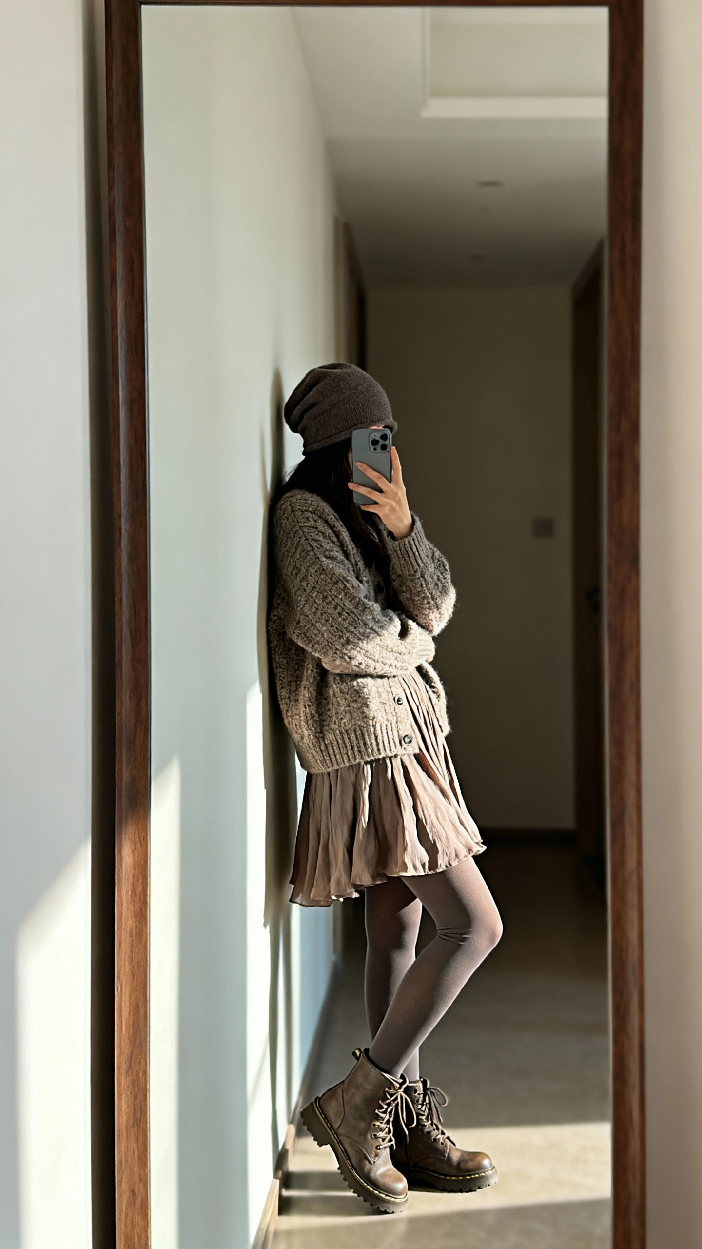 iPhone selfie side-angle in a hallway mirror of a woman wearing a chunky cardigan over a flowy skirt with opaque tights, lace-up boots, and a slouchy beanie, face not visible, soft indoor lighting, casual iPhone aesthetic
