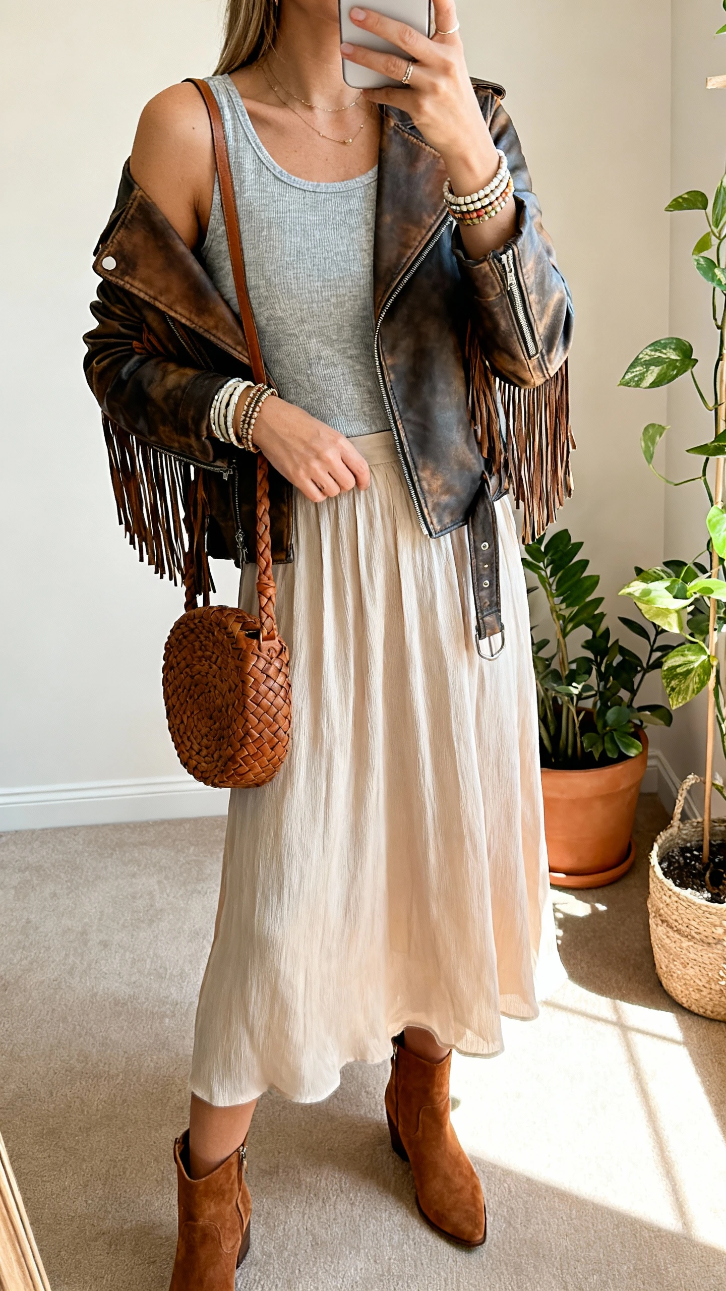 iPhone shoulder-cropped selfie of a woman wearing a mixed-leather jacket with fringe over a soft jersey tee, flowing midi skirt, stacked bangles, suede ankle boots, and a braided leather bag, face not visible, studio corner with plants and warm indoor light, natural iPhone aesthetic.