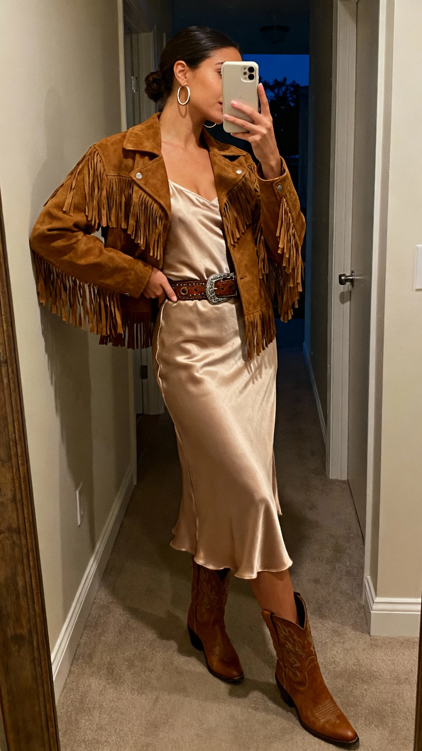 iPhone side-angle mirror selfie of a woman wearing a suede fringe jacket over a satin slip dress, western belt, ankle boots, and hoop earrings, face not visible, hallway mirror with evening indoor lighting, natural iPhone photo quality