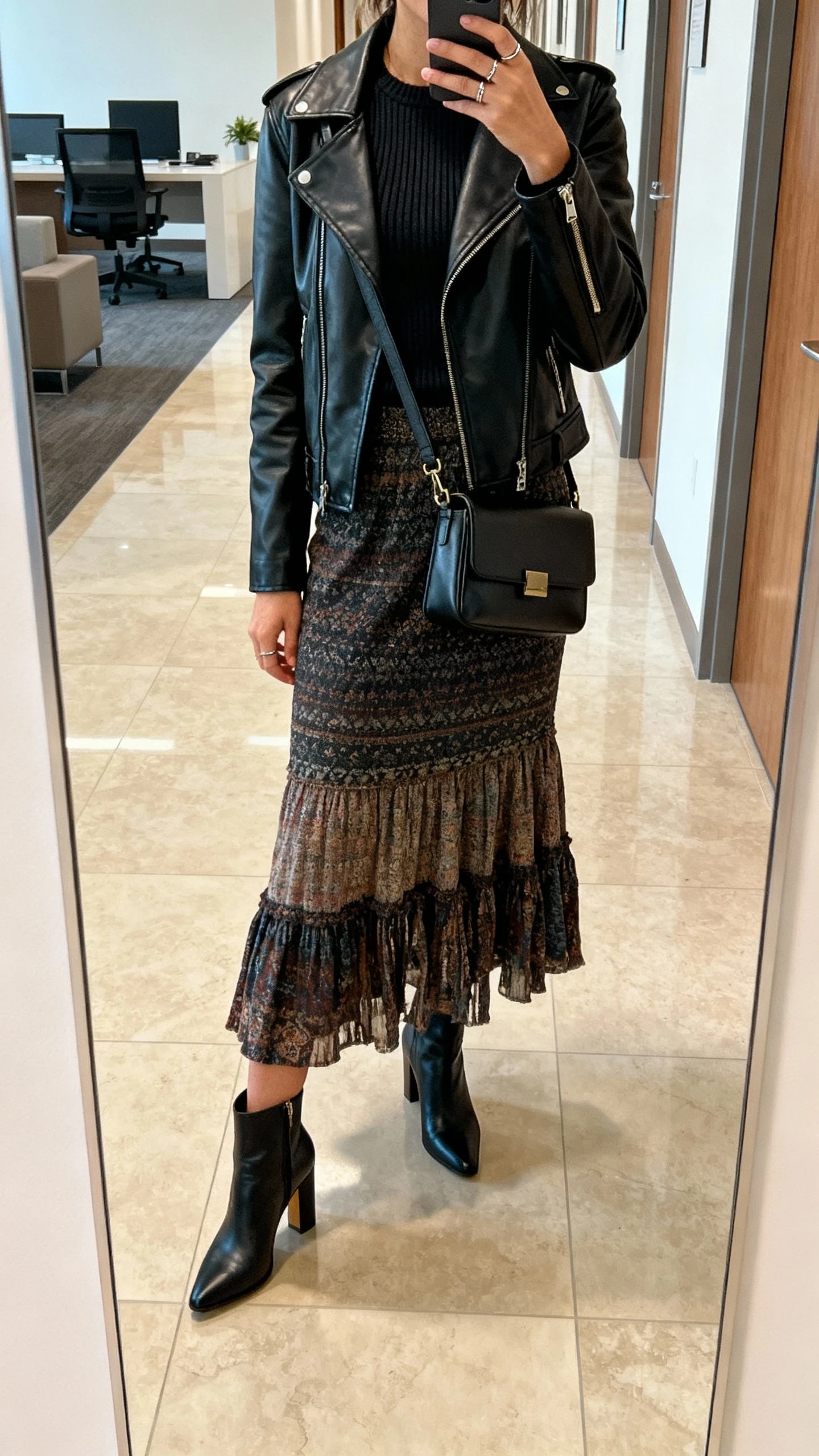 iPhone side-angle mirror selfie of a woman wearing a black leather jacket over a boho skirt, fitted knit top, heeled ankle boots, structured crossbody bag, and minimal rings, face not visible, hallway mirror in an office lobby, natural indoor lighting, casual iPhone shot.