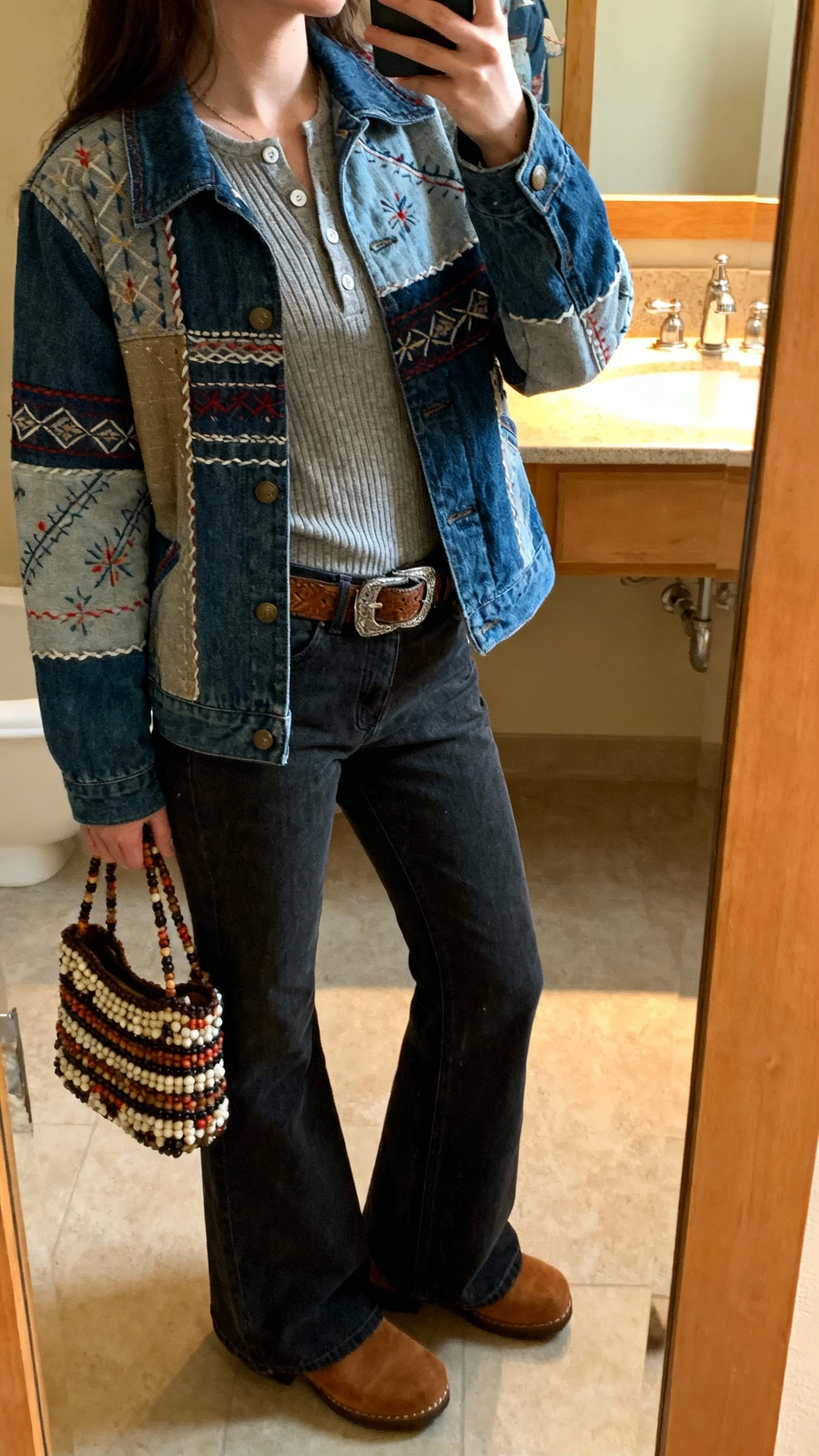iPhone side-angle mirror selfie of a woman wearing a patchwork denim jacket with folky stitching, ribbed henley, flared dark denim, western belt, suede clogs, and a beaded tote, face not visible, bathroom mirror with warm indoor light, natural iPhone aesthetic.