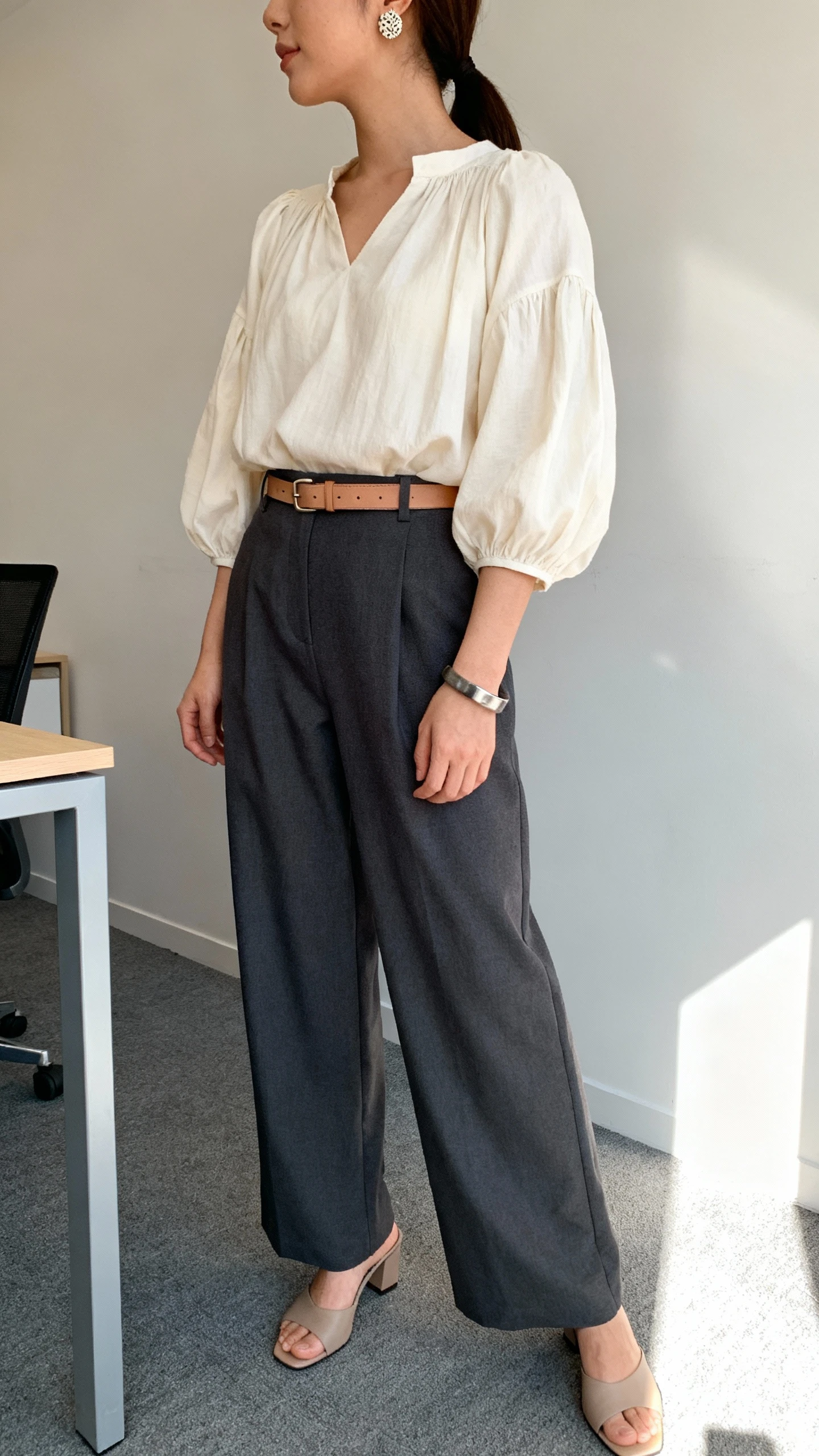 iPhone side-angle office selfie of a woman in a cream billowy peasant blouse tucked into high-waisted charcoal wide-leg trousers, minimal heeled mules, a thin leather belt with subtle hardware, textured stud earrings, and a cuff bracelet, face not visible, morning office light, casual iPhone aesthetic.
