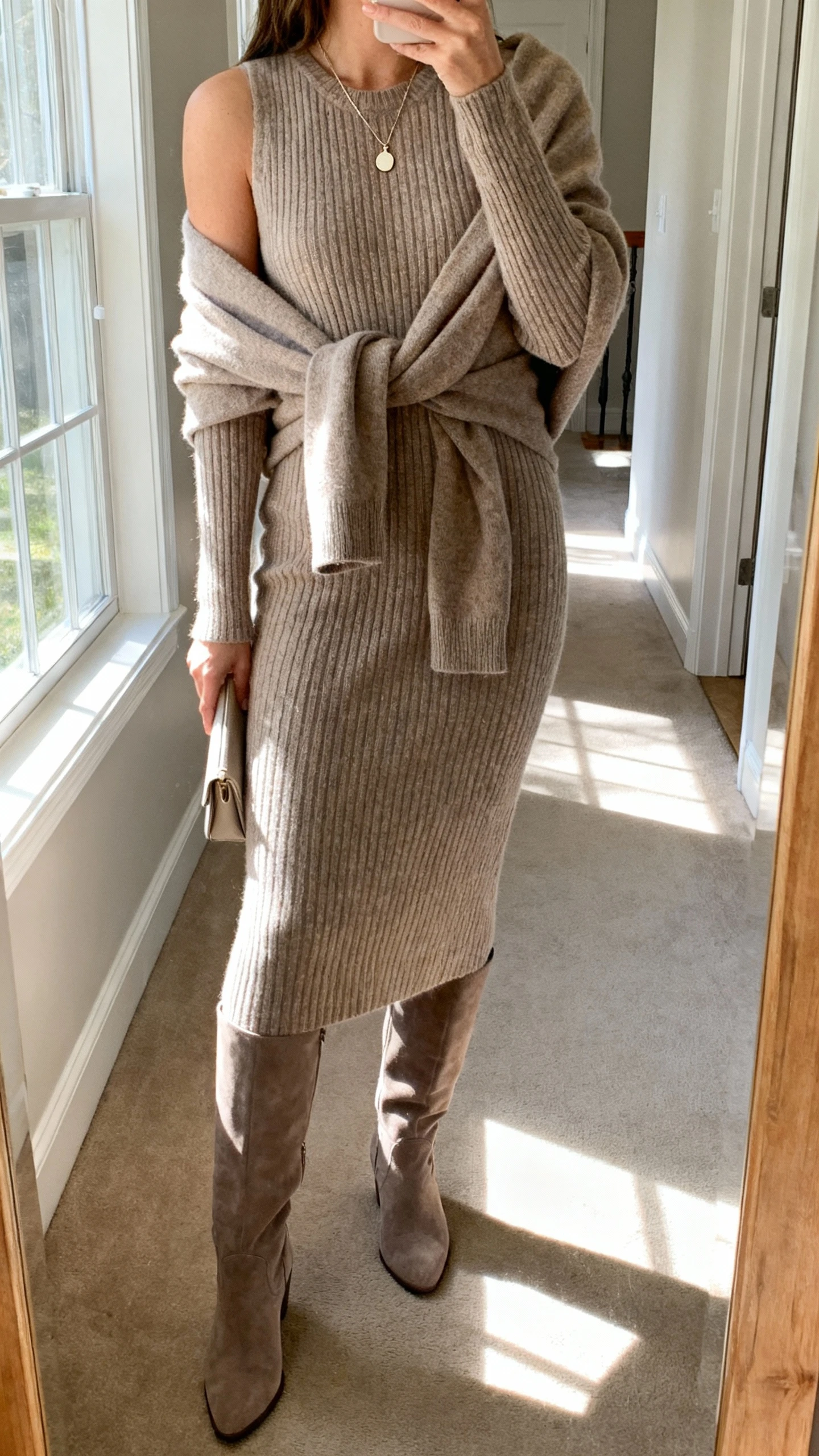 iPhone side-angle selfie cropped at shoulders showing a woman wearing a matching knit sweaterdress set: midi ribbed sweaterdress with a slouchy cardigan tied over shoulders, taupe knee-high boots, delicate pendant necklace, and a minimal clutch, face not visible, hallway mirror at home, afternoon window light, natural iPhone photo.