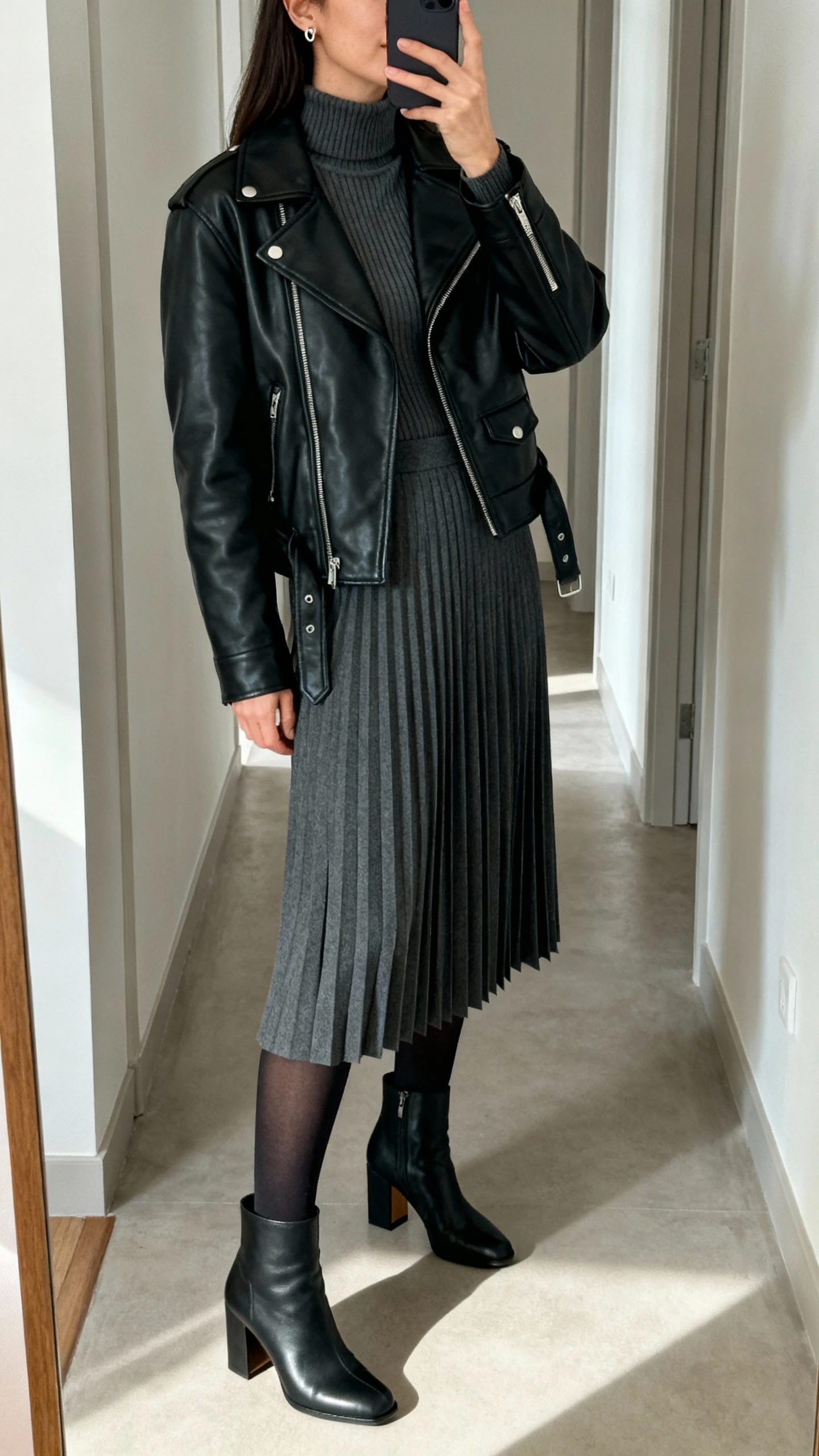 iPhone side-angle selfie in a hallway mirror of a woman wearing a black leather moto jacket over a fitted ribbed turtleneck, charcoal knife-pleated midi skirt, block-heel ankle boots, opaque black tights, and minimal stud earrings; face not visible, natural indoor lighting, casual iPhone photo.