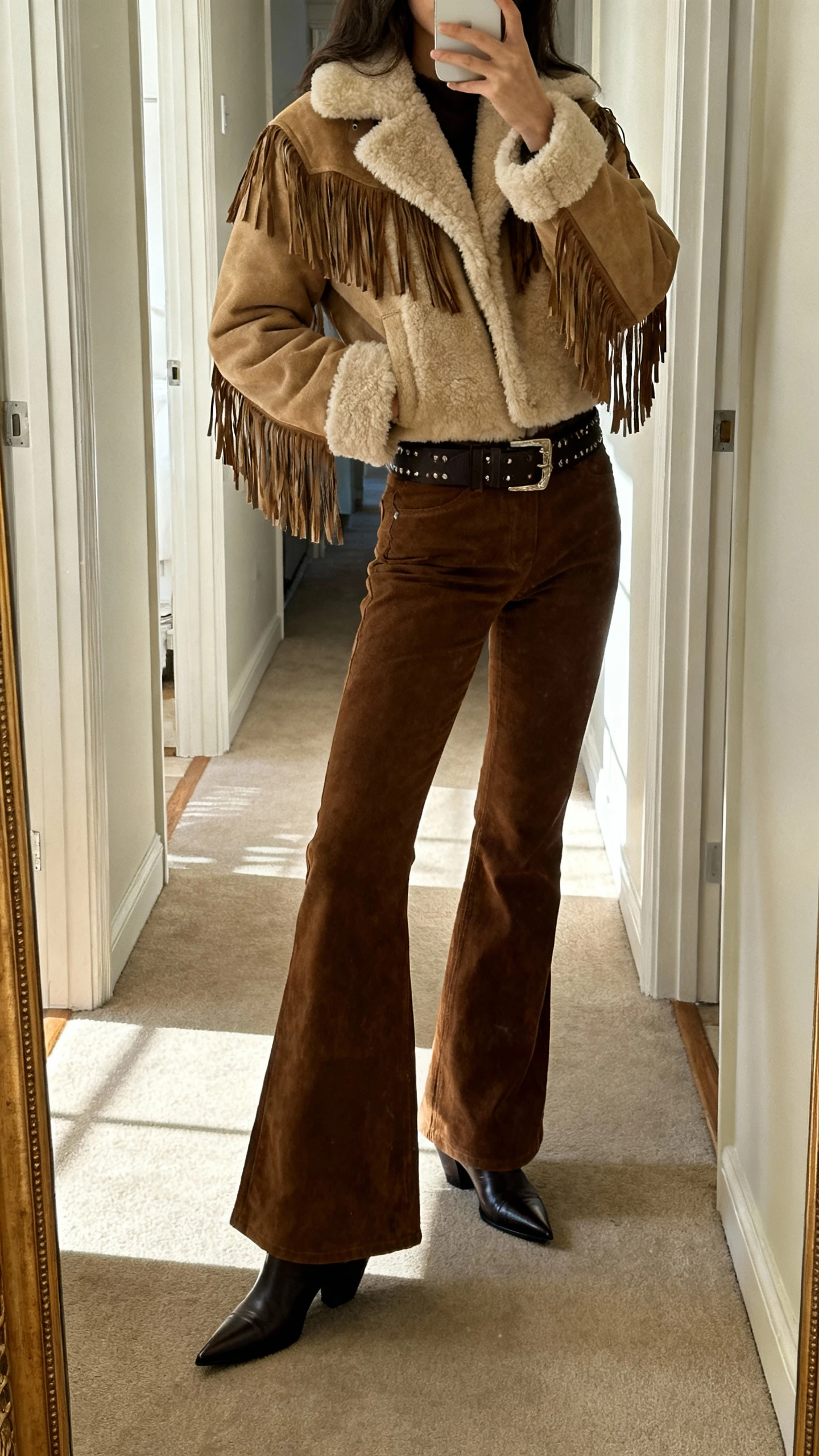 iPhone side-angle selfie in a hallway mirror showing a woman in a shearling jacket with fringe details, retro flares, pointed boots, and a wide belt, face not visible, natural afternoon light, casual iPhone photo.