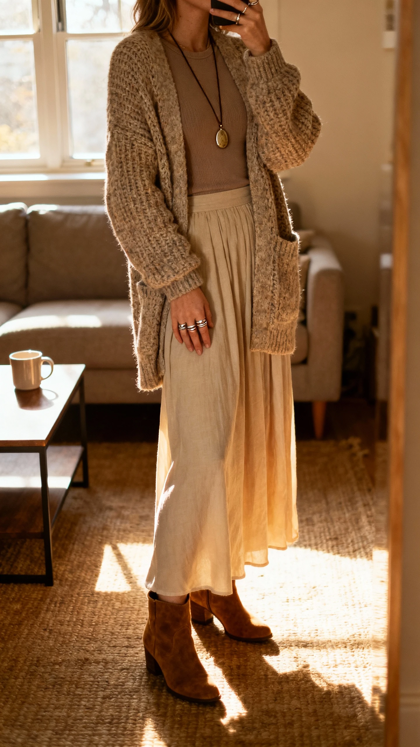 iPhone side-angle selfie of a woman in a chunky cardigan over a fitted top, flowy maxi skirt, suede ankle boots, long pendant necklace, and stacked rings, face not visible, cozy living room by a window with a mug on a coffee table, warm afternoon light, natural iPhone photo.