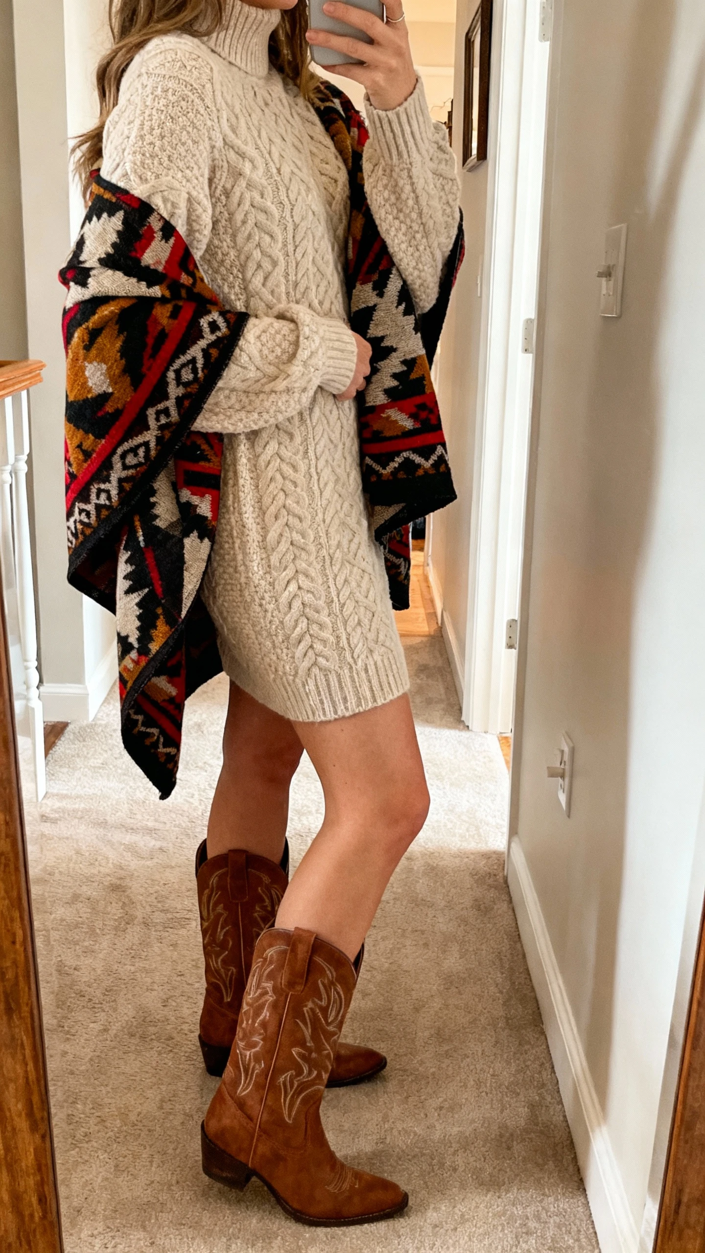 iPhone side-angle selfie of a woman in a cozy cable-knit sweater dress with western boots and a bold patterned statement shawl draped over her shoulders, face not visible, hallway mirror at home, warm indoor lighting, natural iPhone aesthetic.