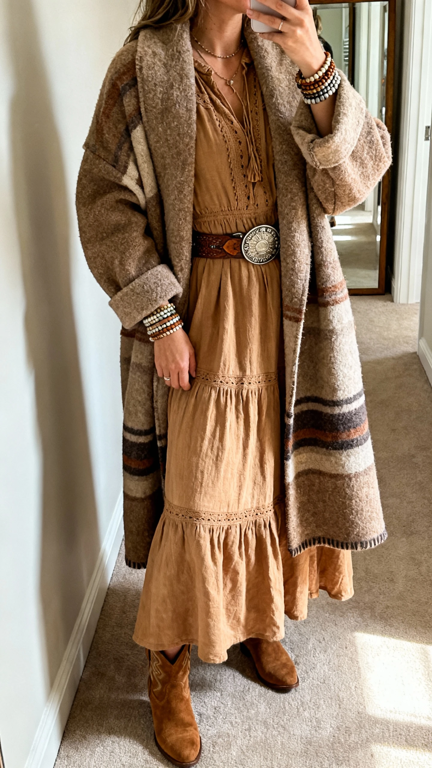iPhone side-angle selfie of a woman in a desert-toned boho maxi dress under a thick blanket coat, stacked bracelets, concho belt, and suede booties; face not visible, hallway mirror, morning light, casual iPhone aesthetic.