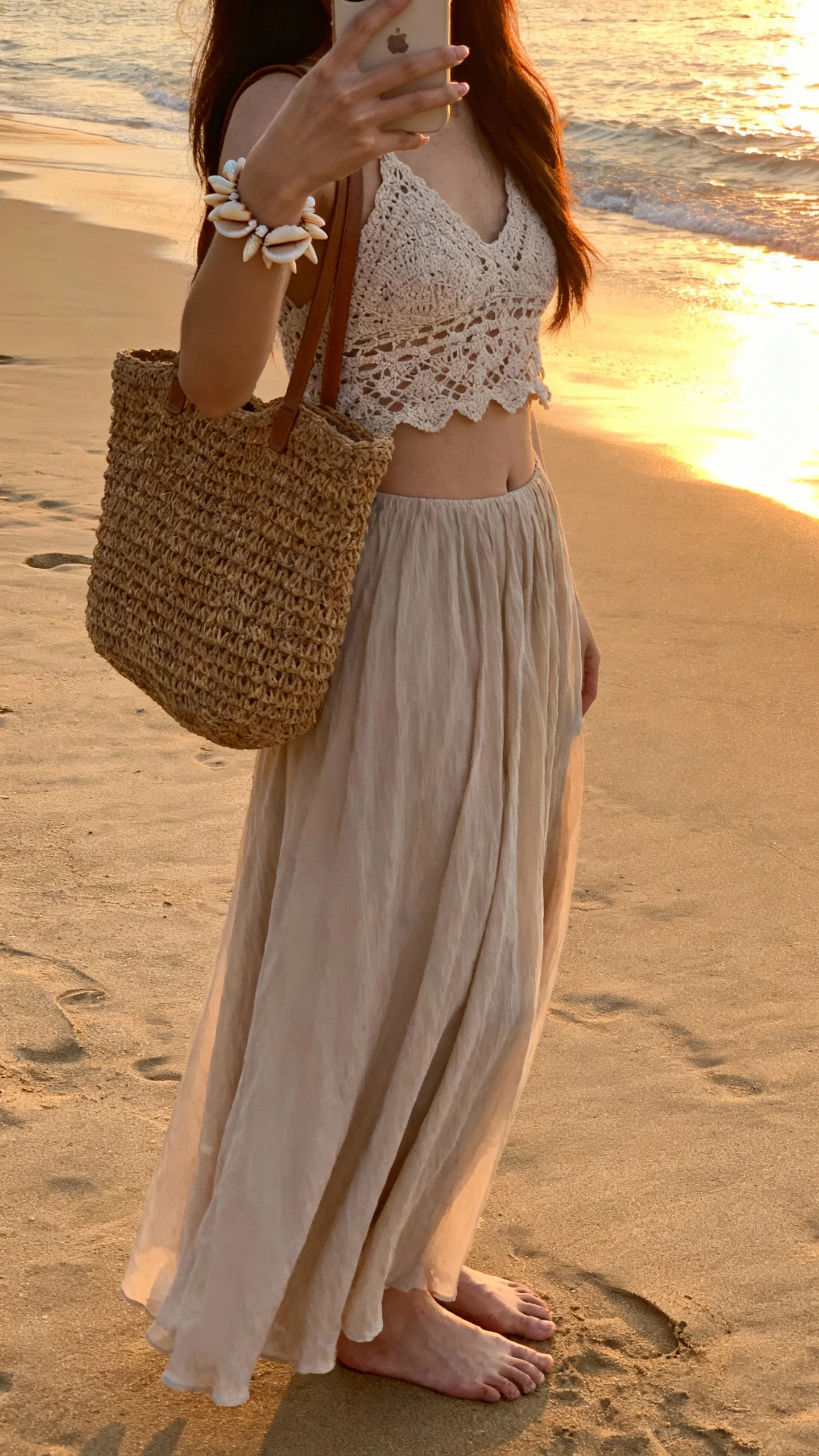iPhone side-angle selfie of a woman in a flowy maxi skirt and a crochet top with shell bracelet and woven tote, standing barefoot on a sandy beach at sunset, face not visible, golden natural light, casual iPhone aesthetic.