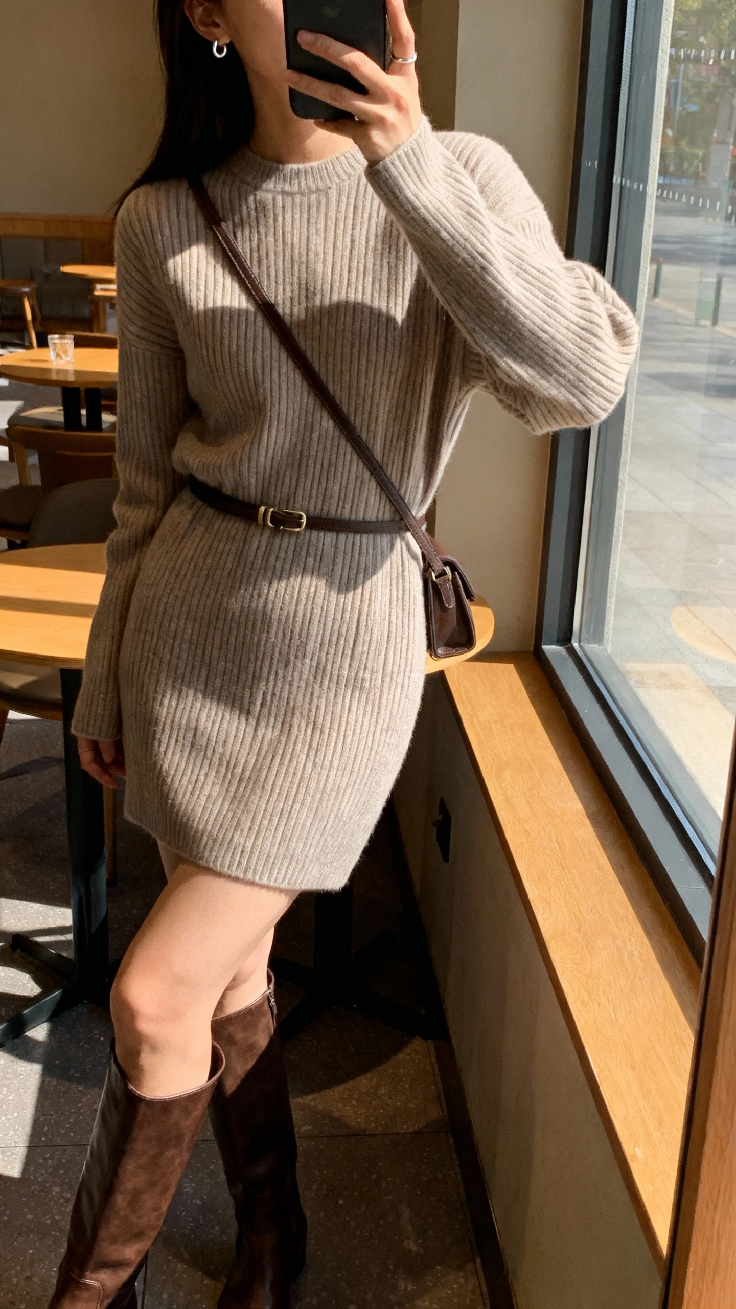 iPhone side-angle selfie of a woman in a ribbed sweater dress with knee-high leather boots, thin waist belt, simple stud earrings, crossbody bag, face not visible, cozy cafe setting with window light, natural iPhone aesthetic.