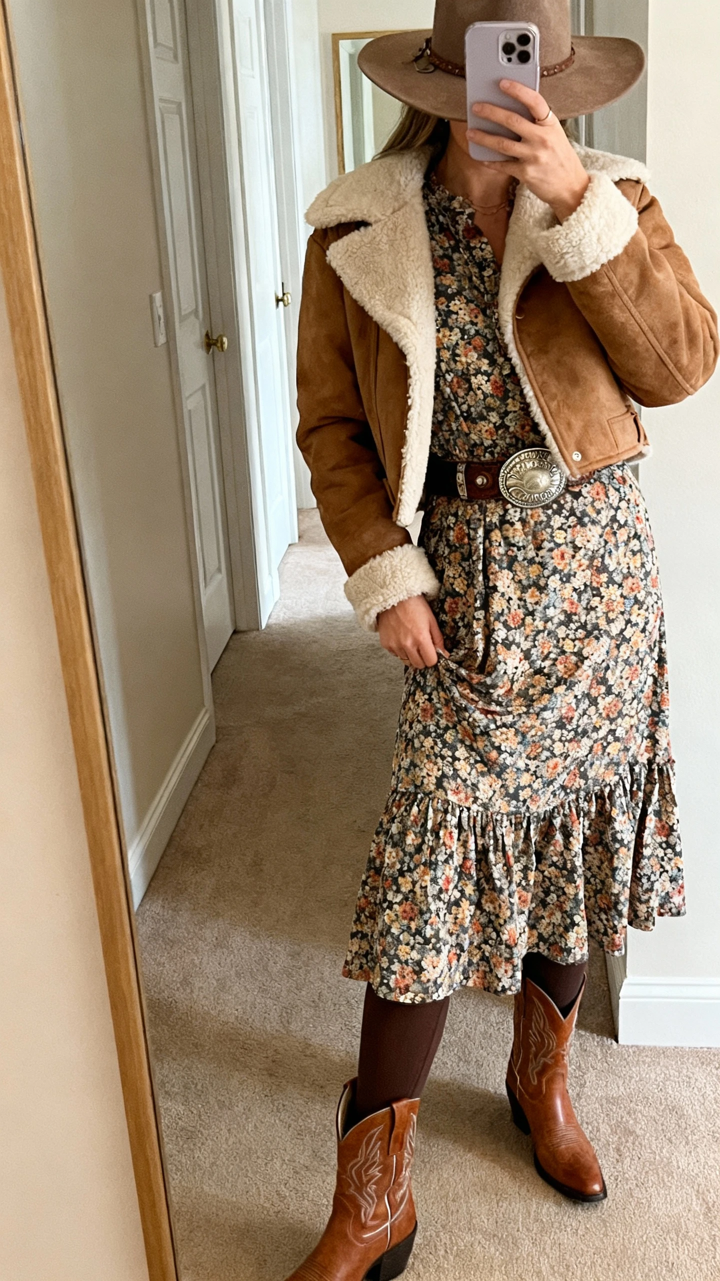 iPhone side-angle selfie of a woman in a shearling shacket layered over a floral midi dress, tights, western ankle boots, and a felt hat with a concho belt, face not visible, hallway mirror with warm indoor light, natural iPhone photo quality.