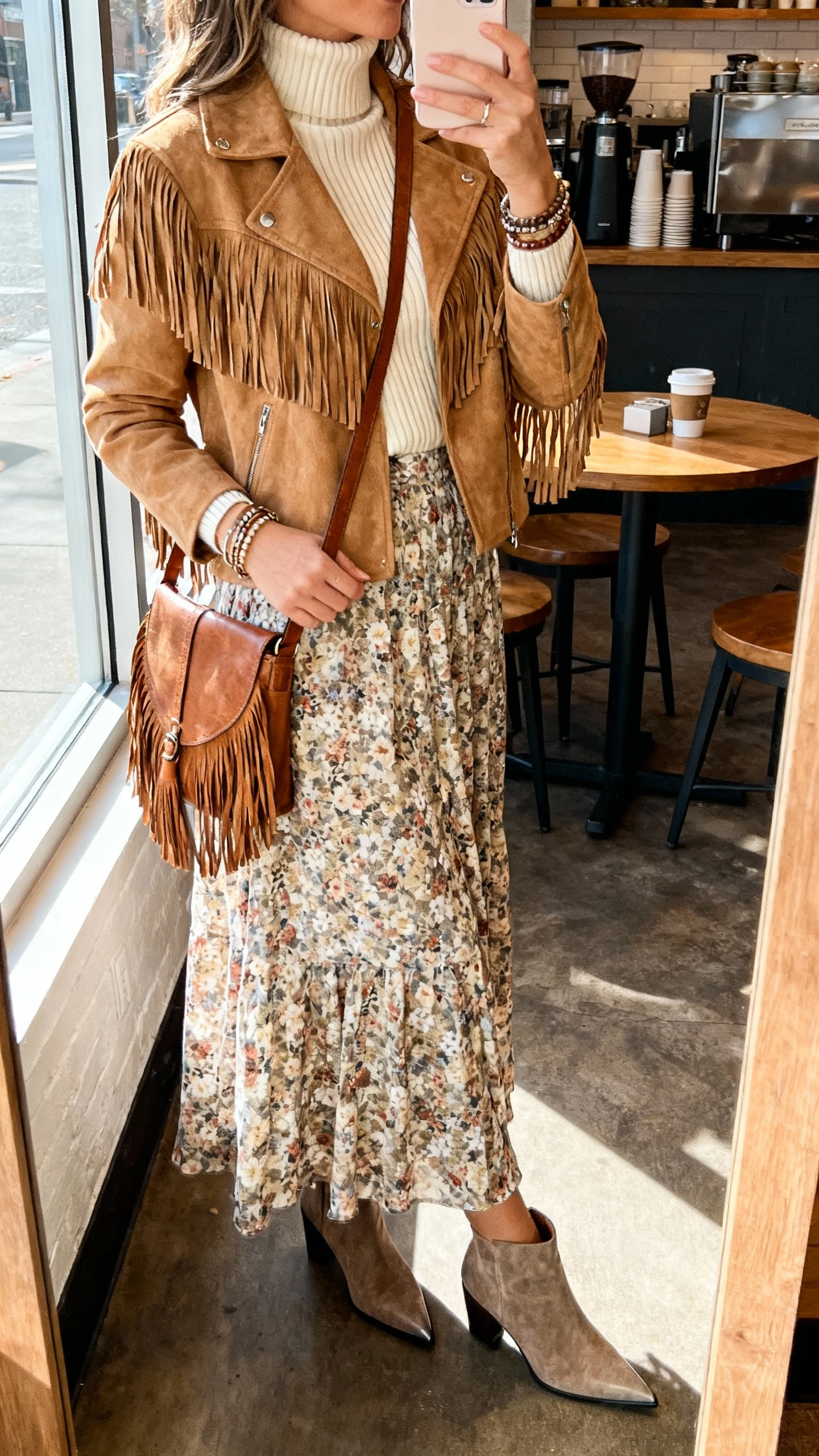 iPhone side-angle selfie of a woman in a tan suede fringe jacket over a cream ribbed turtleneck, floral maxi skirt, pointed-toe ankle boots, leather crossbody with fringe, stacked bracelets, face not visible, taken in a coffee shop near a window, natural afternoon light, iPhone photo quality.