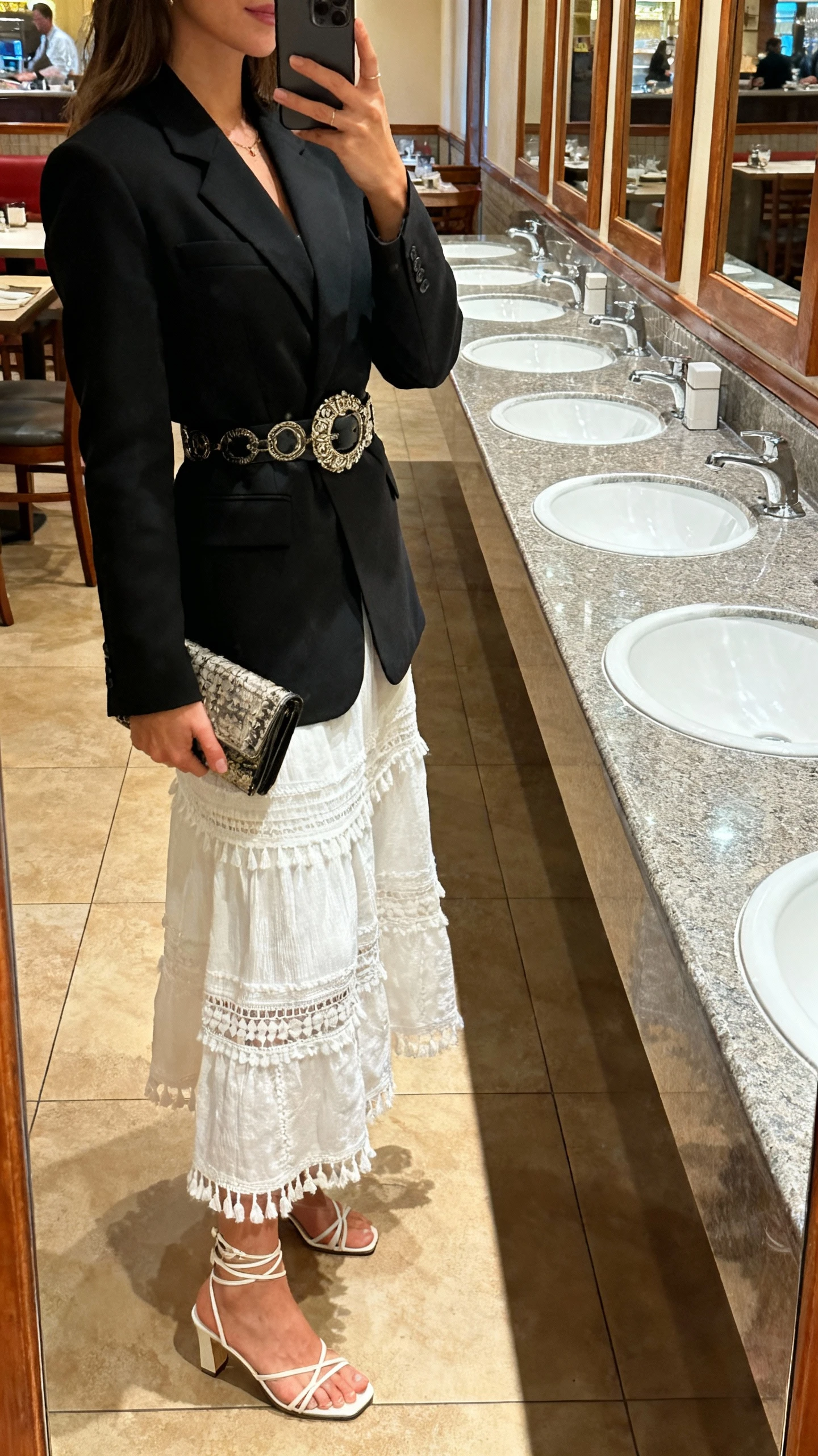 iPhone side-angle selfie of a woman in a white boho skirt, tailored black blazer, cinched with a statement belt, strappy heels, clutch bag, face not visible, restaurant restroom mirror, warm indoor lighting, natural iPhone photo.