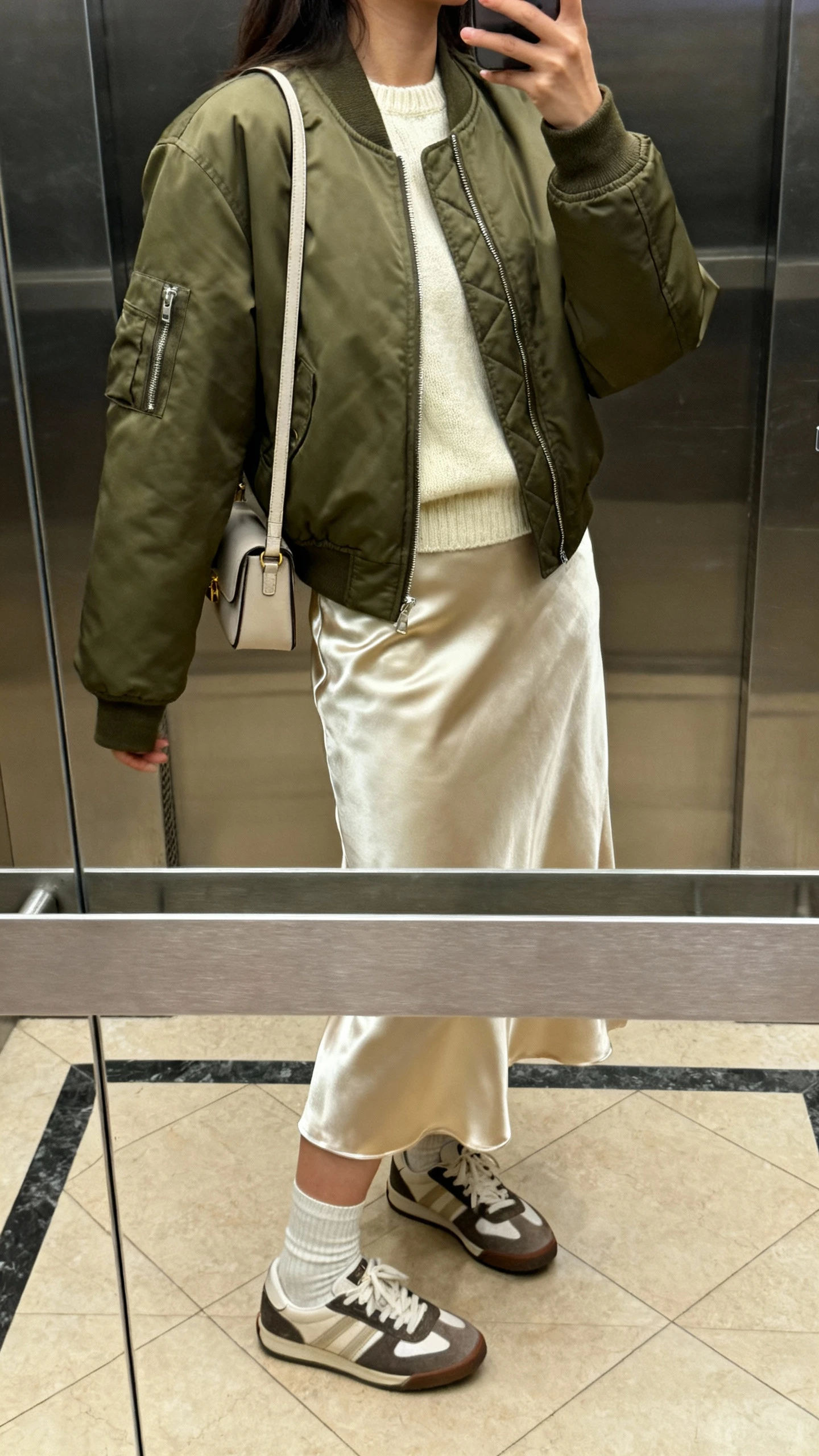 iPhone side-angle selfie of a woman in an olive bomber jacket over a cream sweater, midi satin skirt, ankle socks with retro sneakers, and a small shoulder bag; face not visible, elevator mirror, indoor lighting, natural iPhone aesthetic.