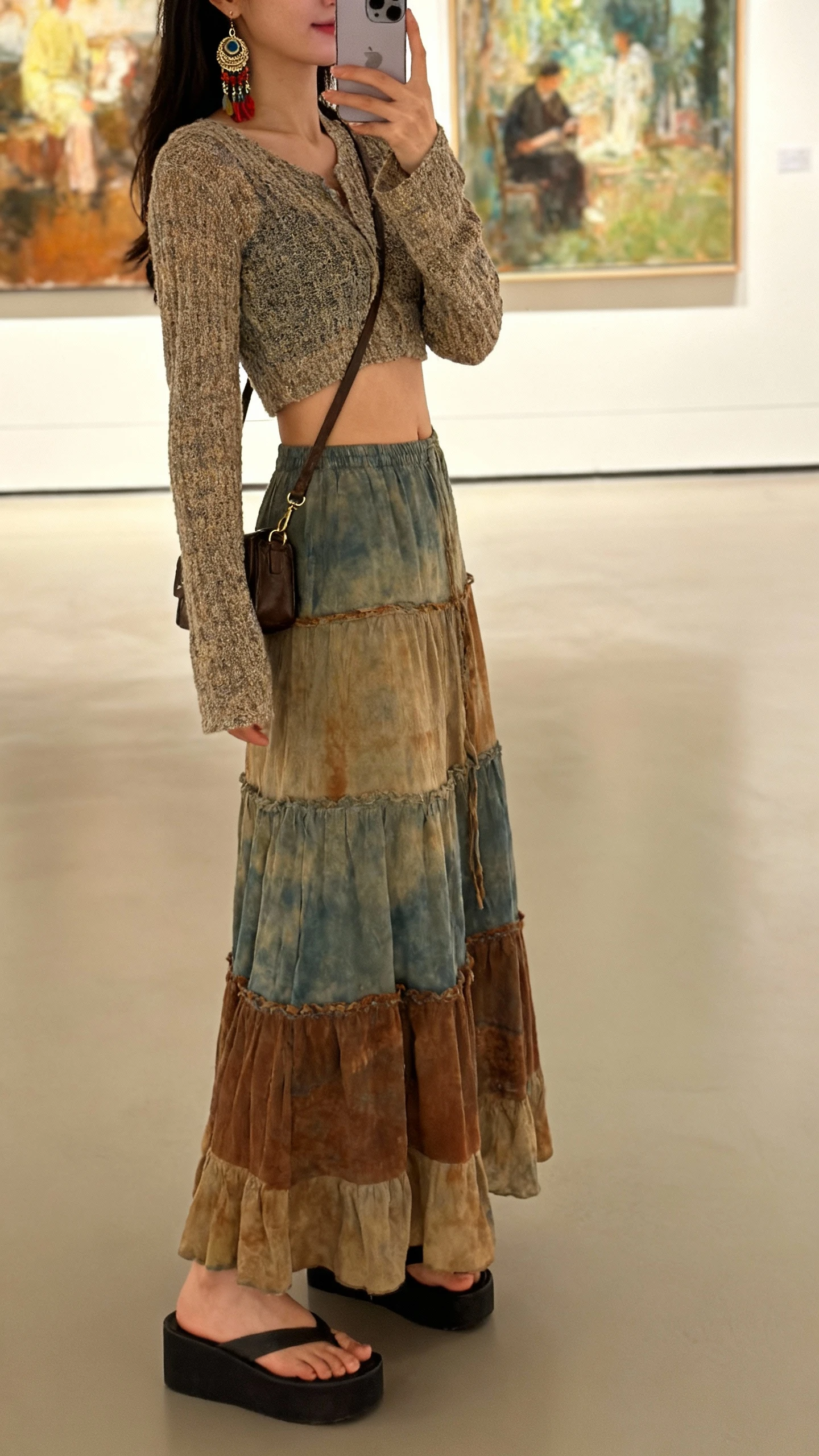 iPhone side-angle selfie of a woman wearing a long boho skirt with a cropped knit top, platform sandals, artsy earrings, small crossbody, face not visible, gallery setting with paintings in background, warm indoor lighting, casual iPhone aesthetic