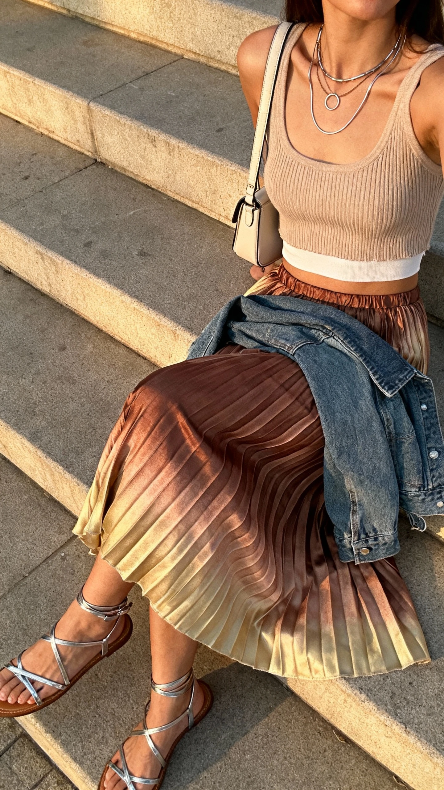 iPhone side-angle selfie seated on steps of a woman in a pleated boho midi skirt (ombre/bronze/soft gold), ribbed knit tank, strappy metallic sandals, mini shoulder bag, mixed-metal layered necklaces with thin hoops, cropped denim jacket draped over lap, face not visible, late-afternoon natural light, casual iPhone aesthetic.