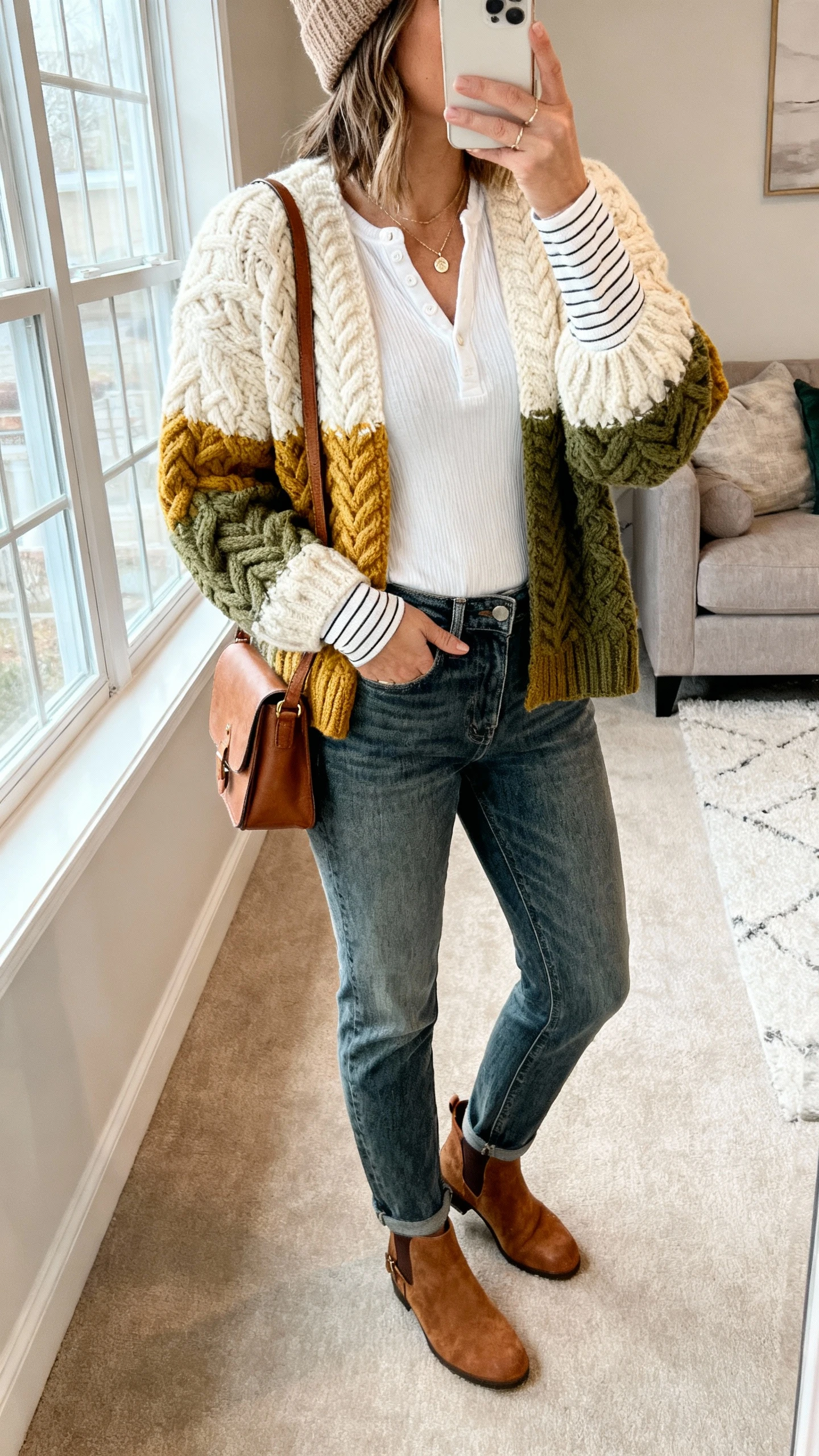 iPhone side-angle selfie showing a woman wearing a chunky cable-knit cardigan (cream/honey/moss), fitted long-sleeve tee or henley (white/striped), high-rise straight jeans (mid-wash), suede ankle booties or leather clogs, optional wool beanie, leather crossbody, dainty pendant, face not visible, cozy living room setting with window light, casual iPhone photo.