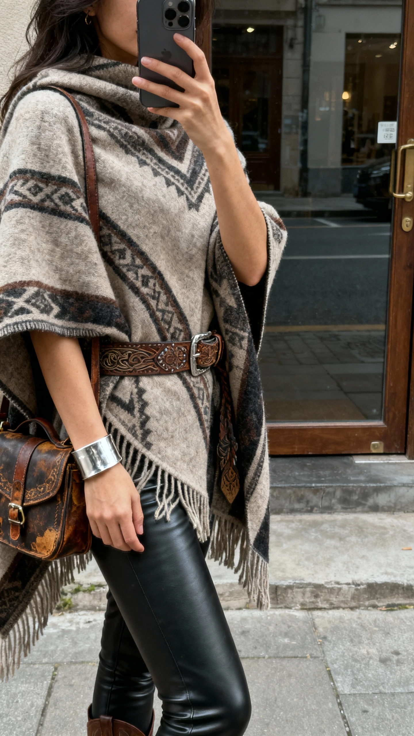 iPhone side-profile selfie of a woman in a statement poncho over leather leggings with heritage accessories (tooled belt, vintage-style bag, and silver cuff), face not visible, street doorway reflection, overcast daylight, natural iPhone photo quality.