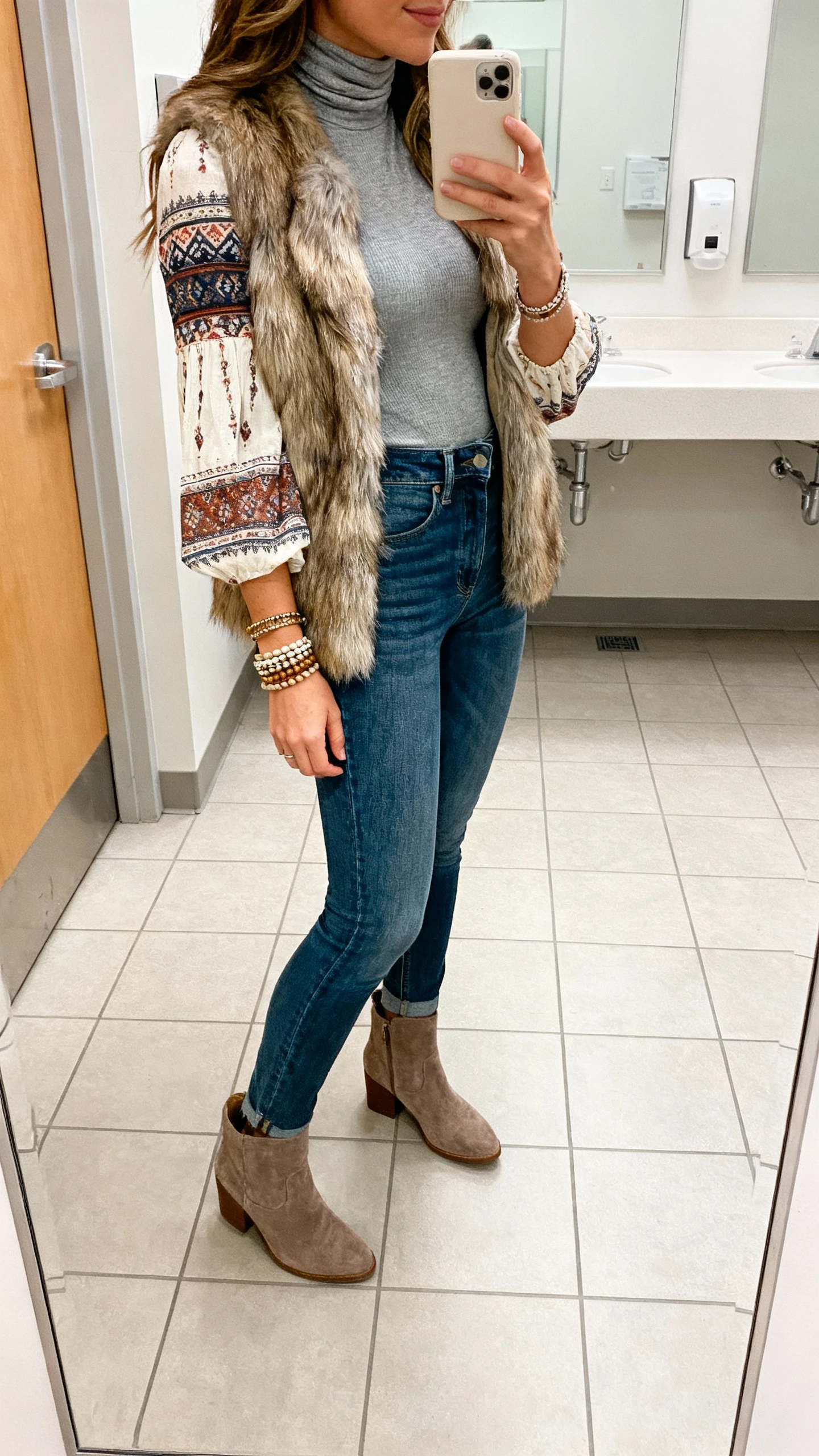 iPhone side-view selfie of a woman wearing a fitted turtleneck layered under a boho blouse, high-rise jeans, and a faux-fur vest with stacked bracelets and suede booties, face not visible, office restroom mirror, bright indoor lighting, casual iPhone photo.