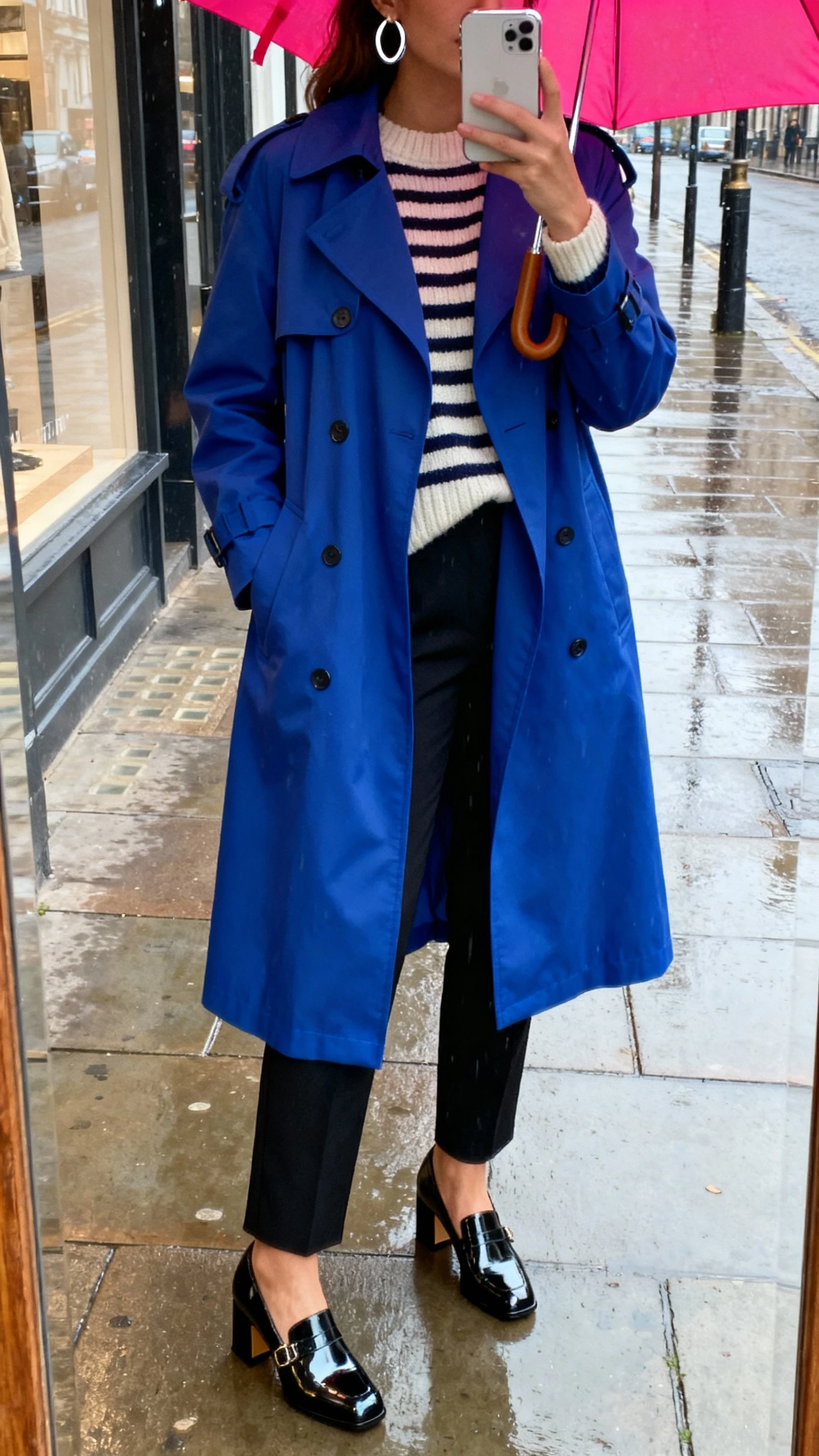 iPhone street-mirror selfie of a woman in a bold color-pop trench coat in cobalt over a striped knit, black cigarette trousers, patent block-heel Mary Janes, bright umbrella, and silver hoop earrings, face not visible, rainy sidewalk storefront mirror, natural overcast light, iPhone photo quality.