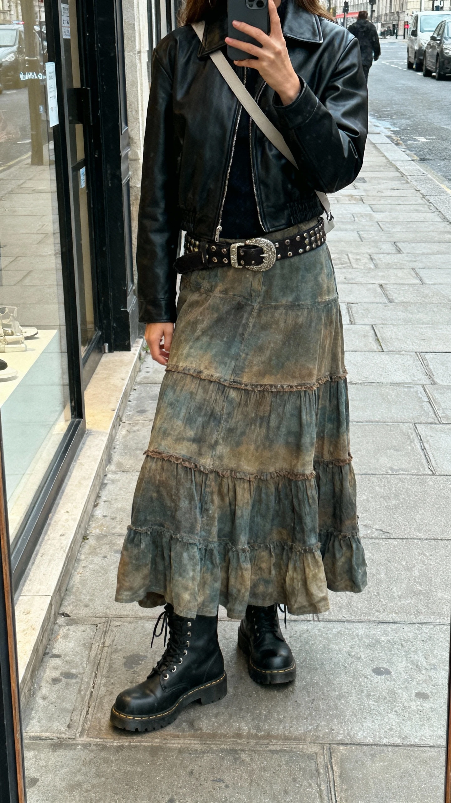 iPhone street mirror selfie (shop window reflection) of a woman wearing a long boho skirt with a black leather jacket, combat boots, chunky belt, minimal crossbody, face not visible, overcast city sidewalk, natural daylight, casual iPhone aesthetic