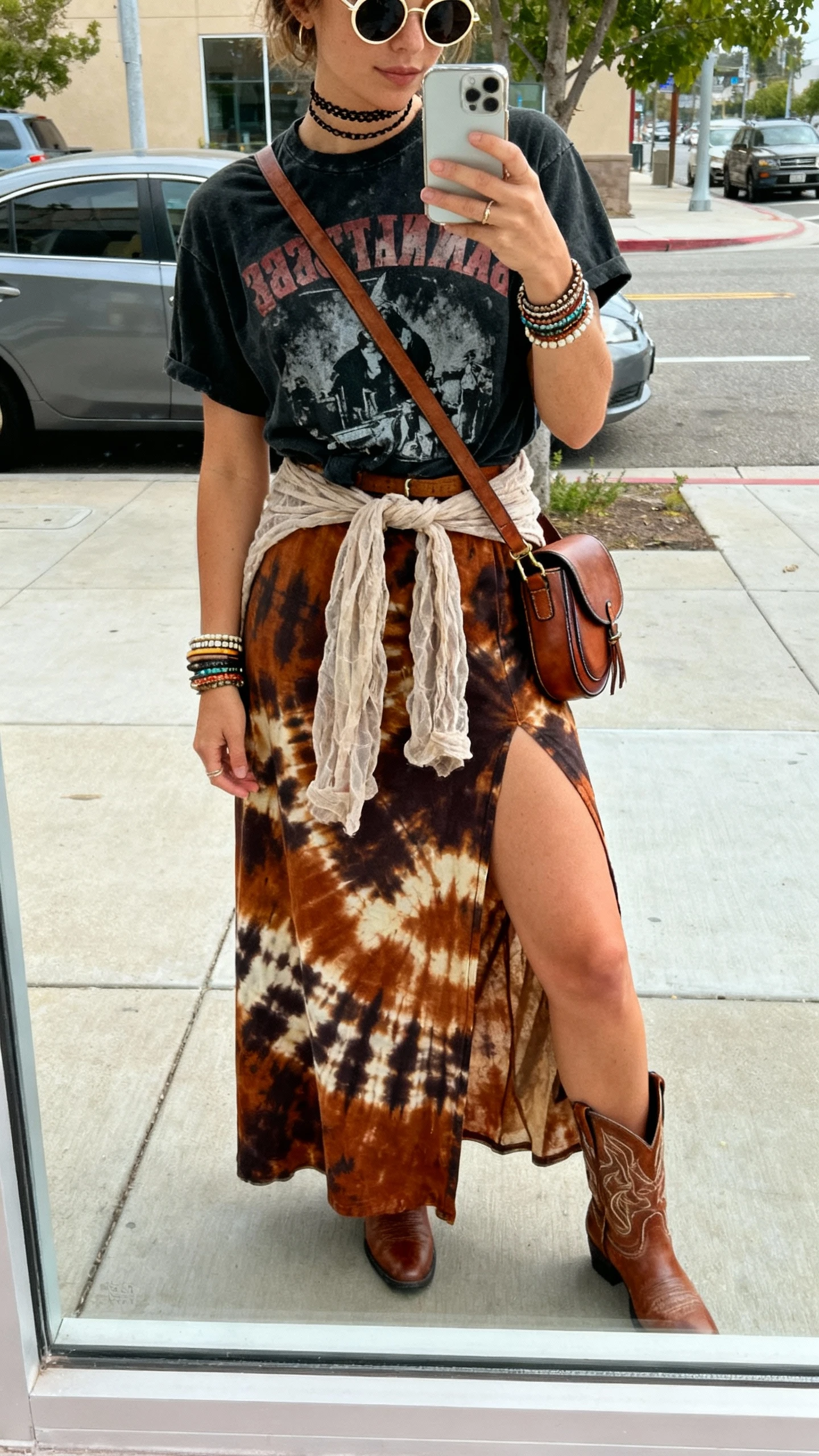 iPhone street mirror window selfie of a woman in a high-slit boho midi/maxi skirt (bold print or earthy tie-dye), washed black graphic band tee tucked, western ankle boots, crossbody saddle bag, stacked bangles and layered chokers, round sunglasses, gauzy duster tied at waist, face not visible, natural daylight, casual iPhone vibe.