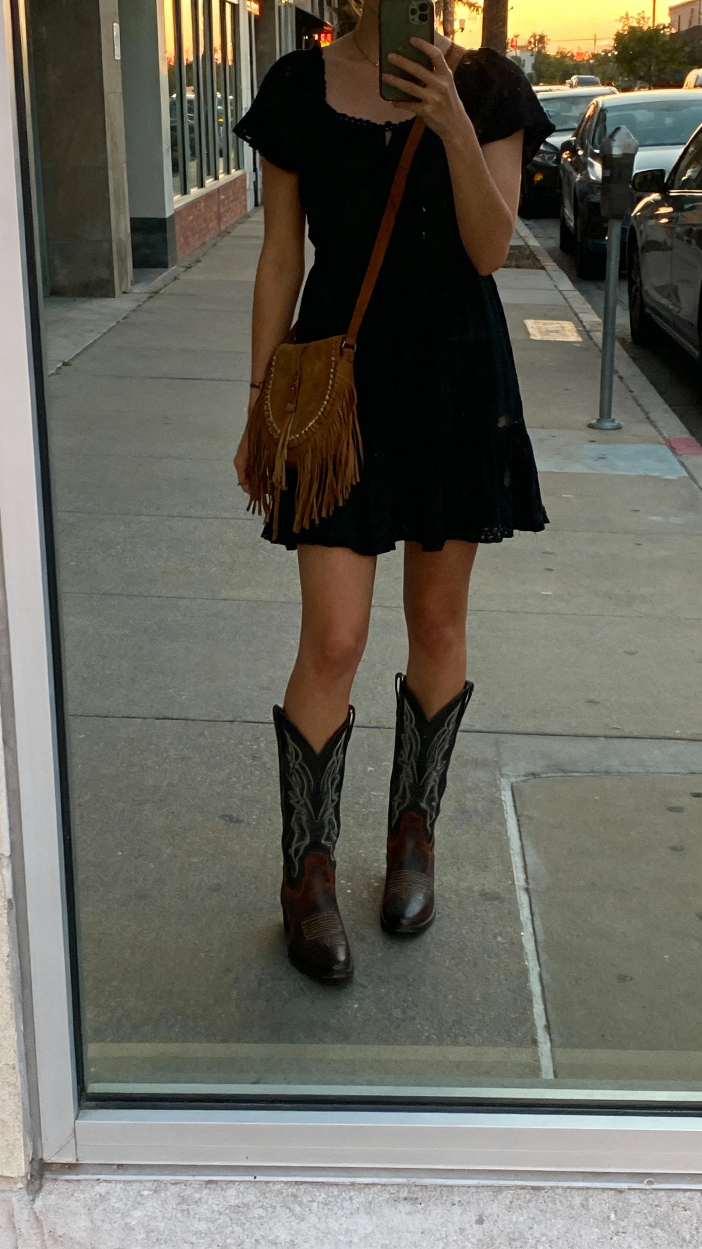 iPhone street mirror window selfie of a woman in a boho little black dress with western-edge boots and a fringe crossbody, face not visible, city sidewalk at dusk, natural ambient light, casual iPhone photo.