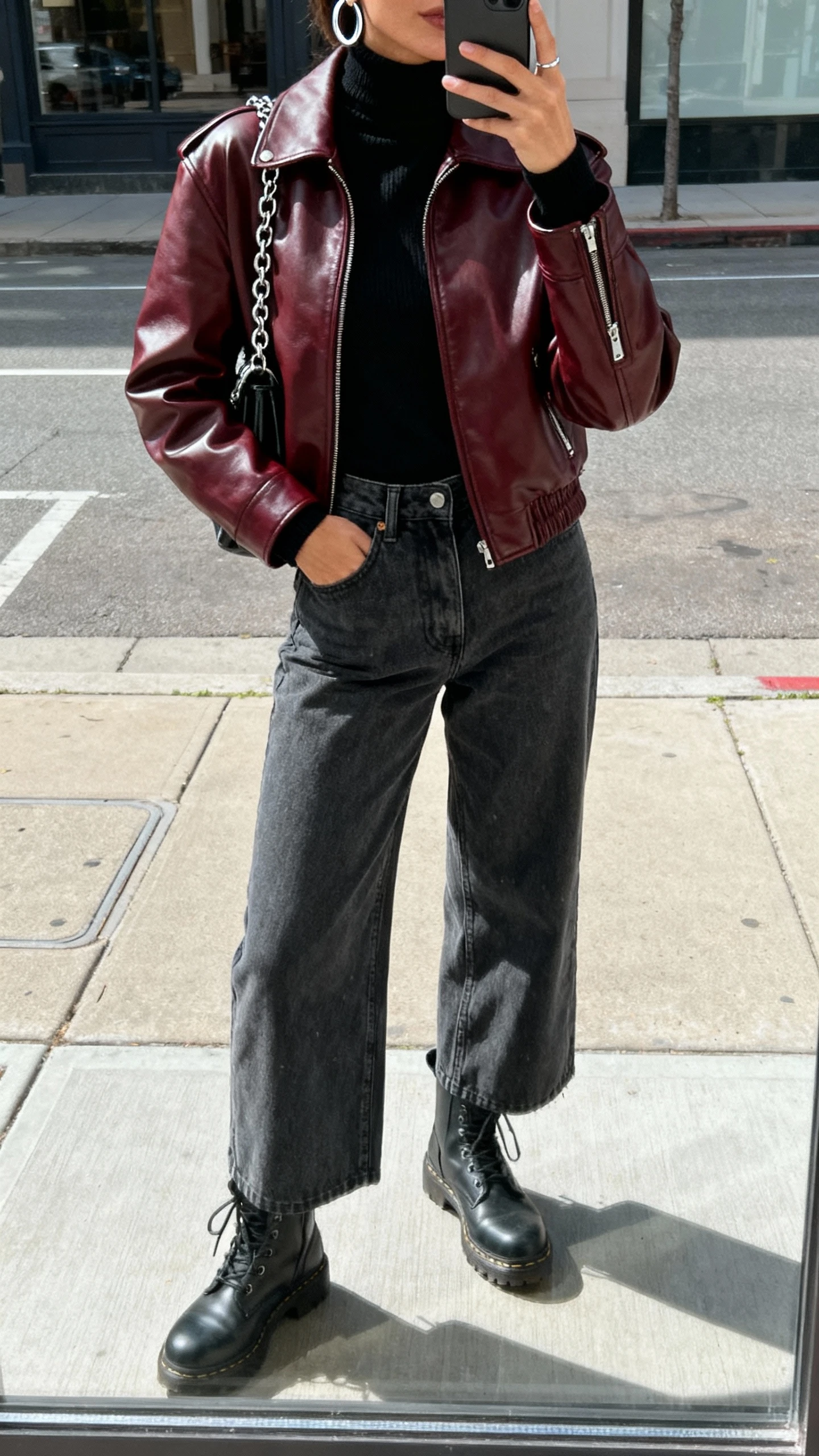iPhone street mirror-window selfie of a woman in a burgundy leather moto jacket layered over a black turtleneck, charcoal wide-leg jeans, combat boots, silver hoops, and a chain-strap bag, face not visible, urban sidewalk reflection in daylight, casual iPhone photo.