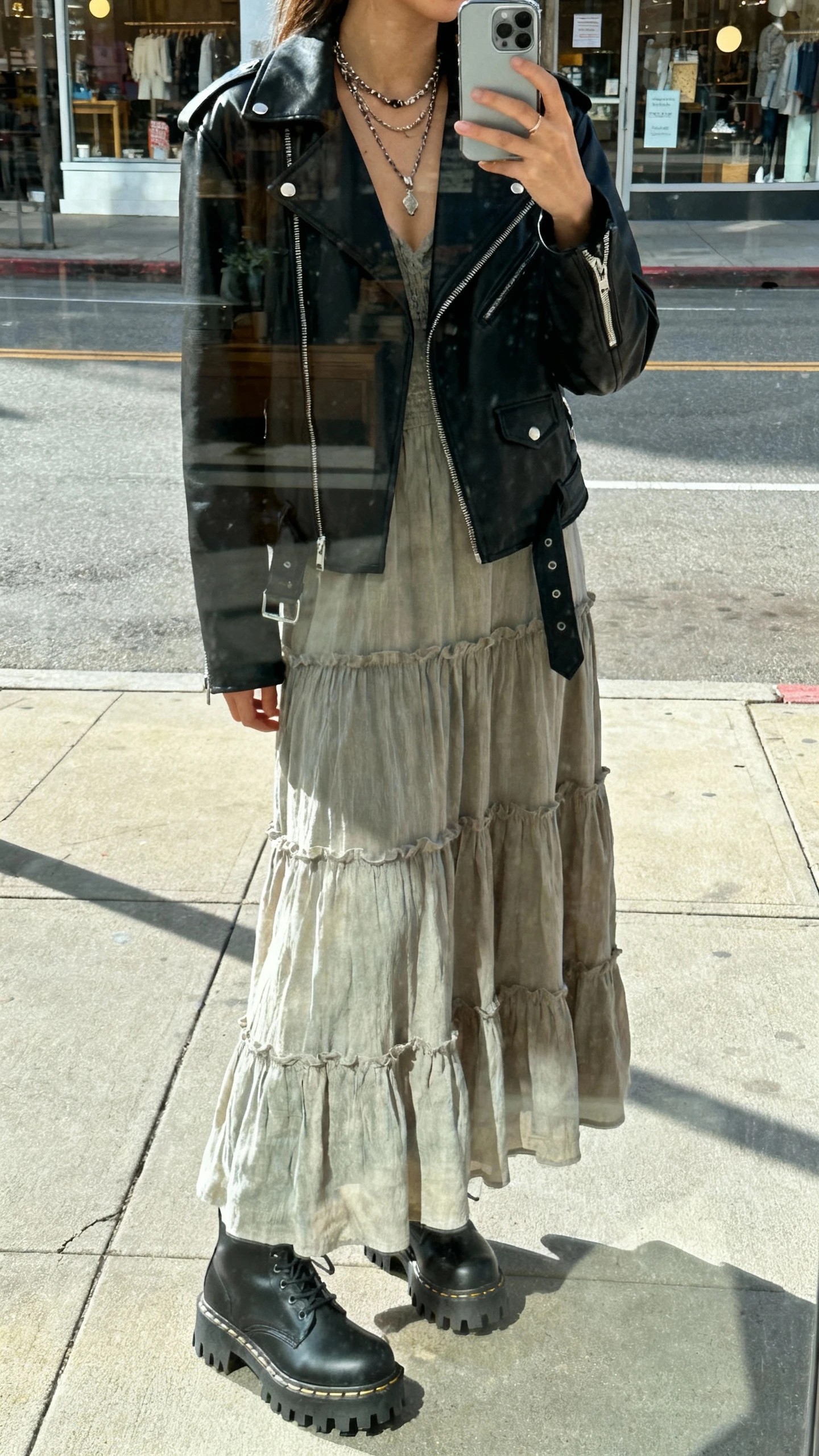 iPhone street reflection selfie showing a woman in a tiered maxi dress under a black moto jacket with lug-sole boots, layered necklaces, face not visible, city sidewalk shop-window reflection, daylight, casual iPhone vibe.