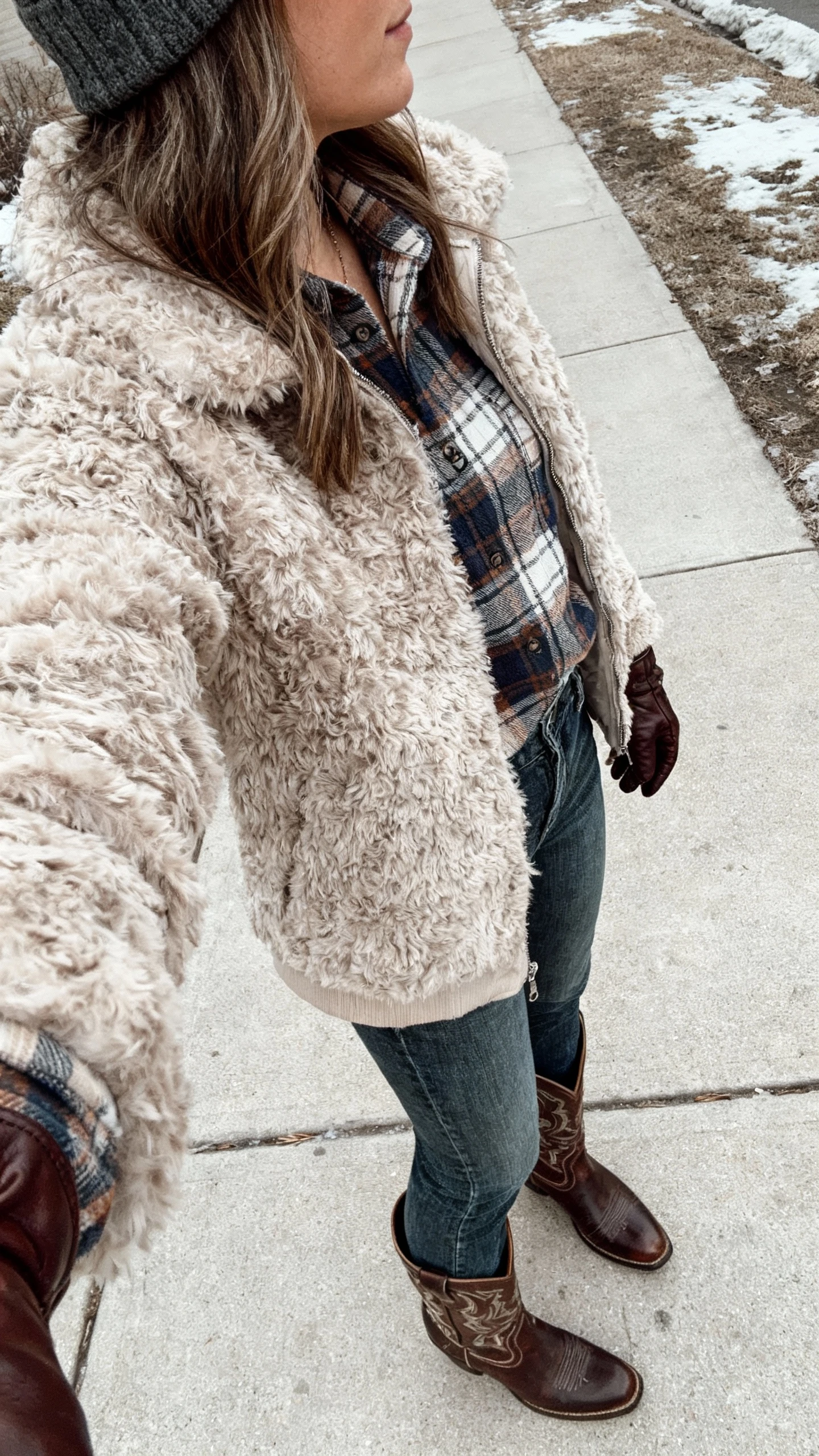 iPhone street-style side-view selfie of a woman in a shaggy faux fur jacket layered over a plaid shirt with straight-leg jeans and classic cowboy boots, plus a beanie and leather gloves, face not visible, chilly sidewalk scene in overcast natural light, natural iPhone photo quality.
