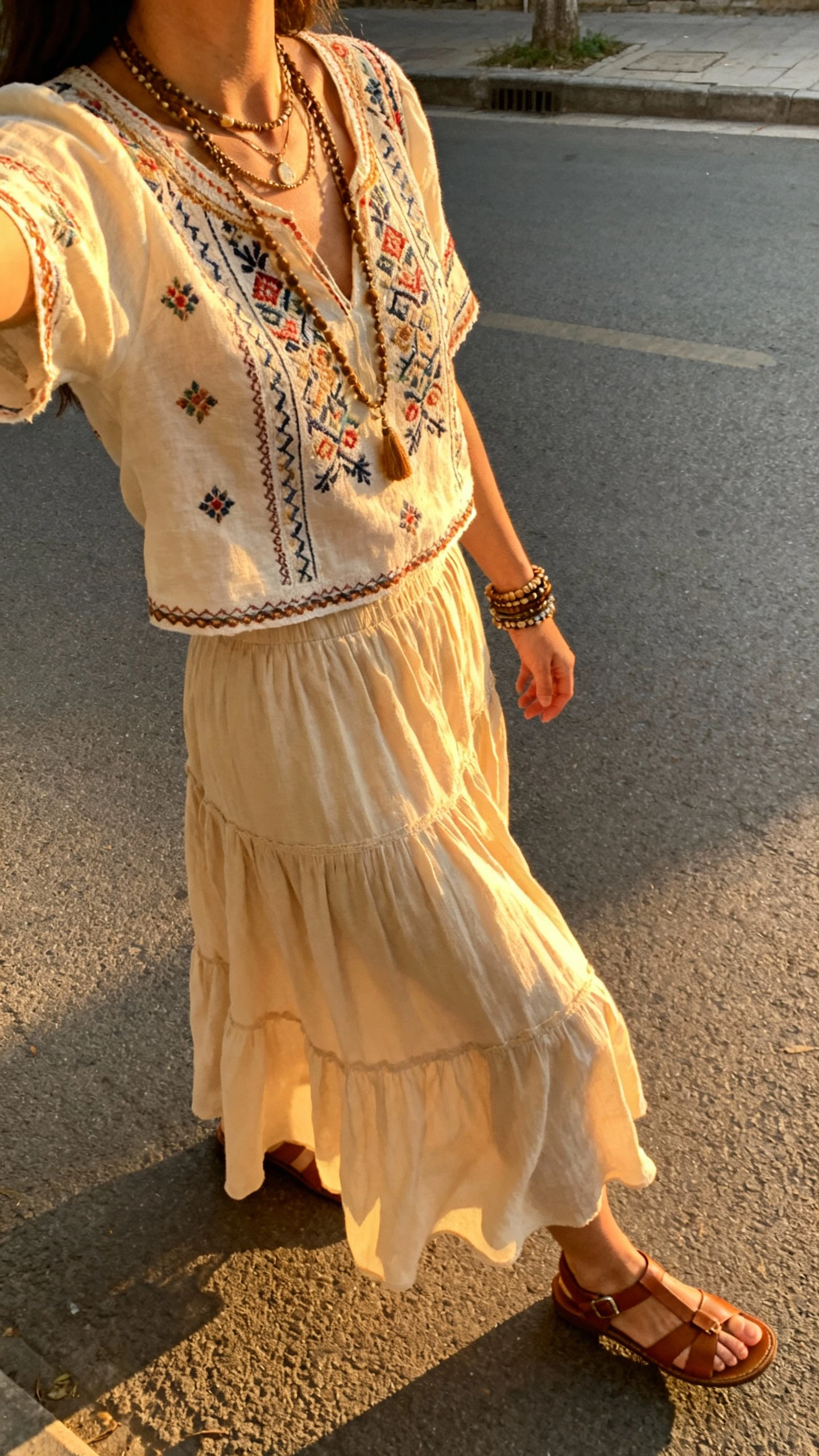 iPhone street-style side view selfie of a woman wearing a flowy skirt, embroidered boho top, layered necklaces and bracelets, leather sandals, face not visible, golden hour light on a quiet street, natural iPhone photo quality.