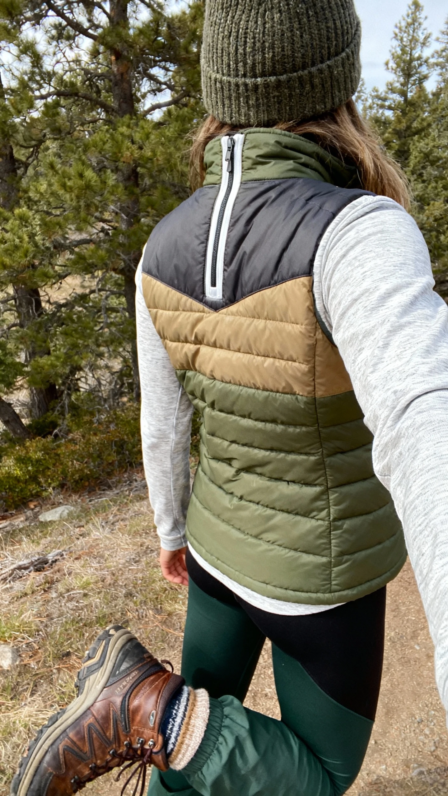 iPhone trailhead back-view selfie of a woman wearing a quilted/puffer vest (olive/charcoal/tan), moisture-wicking long-sleeve or quarter-zip (cream/heather gray), thick leggings or hiking pants (black/deep green), trail sneakers or rugged leather hikers with cozy socks peeking, wool beanie, face not visible, natural daylight by trees, casual iPhone photo.