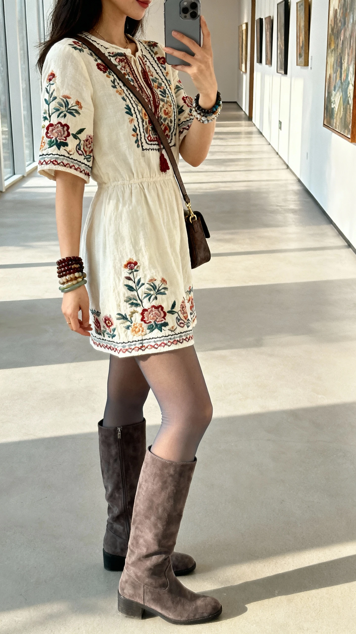 Side-angle iPhone selfie of a woman in an embroidered mini dress with opaque tights and over-the-knee boots, stacked bracelets and a crossbody bag, face not visible, gallery hallway setting, natural daylight, casual iPhone photo.