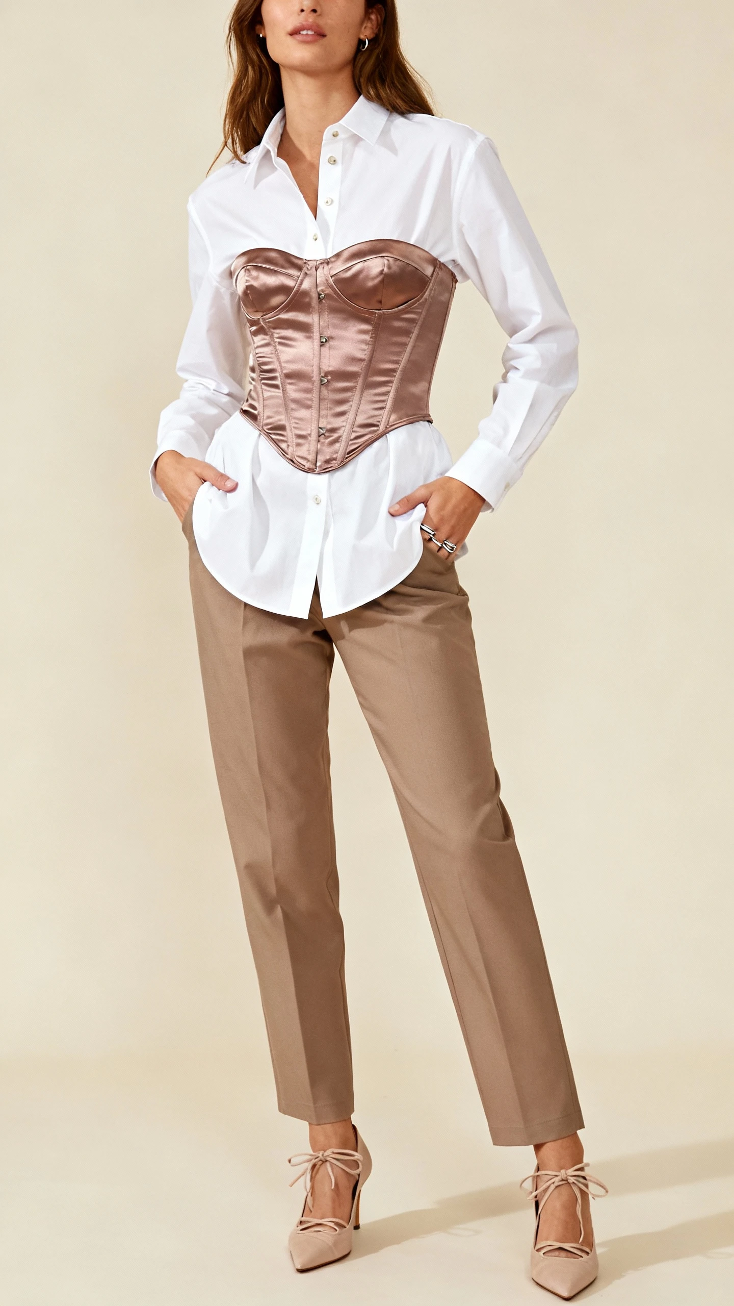 Studio portrait of a woman wearing a satin corset layered over a crisp white button-up shirt, straight-leg trousers, lace-up heels, minimal silver rings, neutral beige background, soft studio lighting, relaxed hands-in-pockets pose, clean composition.