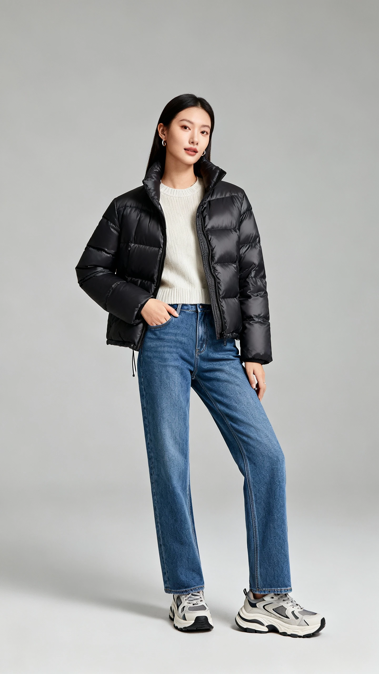 Studio fashion photo of a woman wearing a sleek elevated puffer jacket with straight-leg blue jeans, simple knit top, chunky sneakers, and minimal accessories; neutral gray background, studio lighting, professional natural pose, clean composition, magazine-quality.